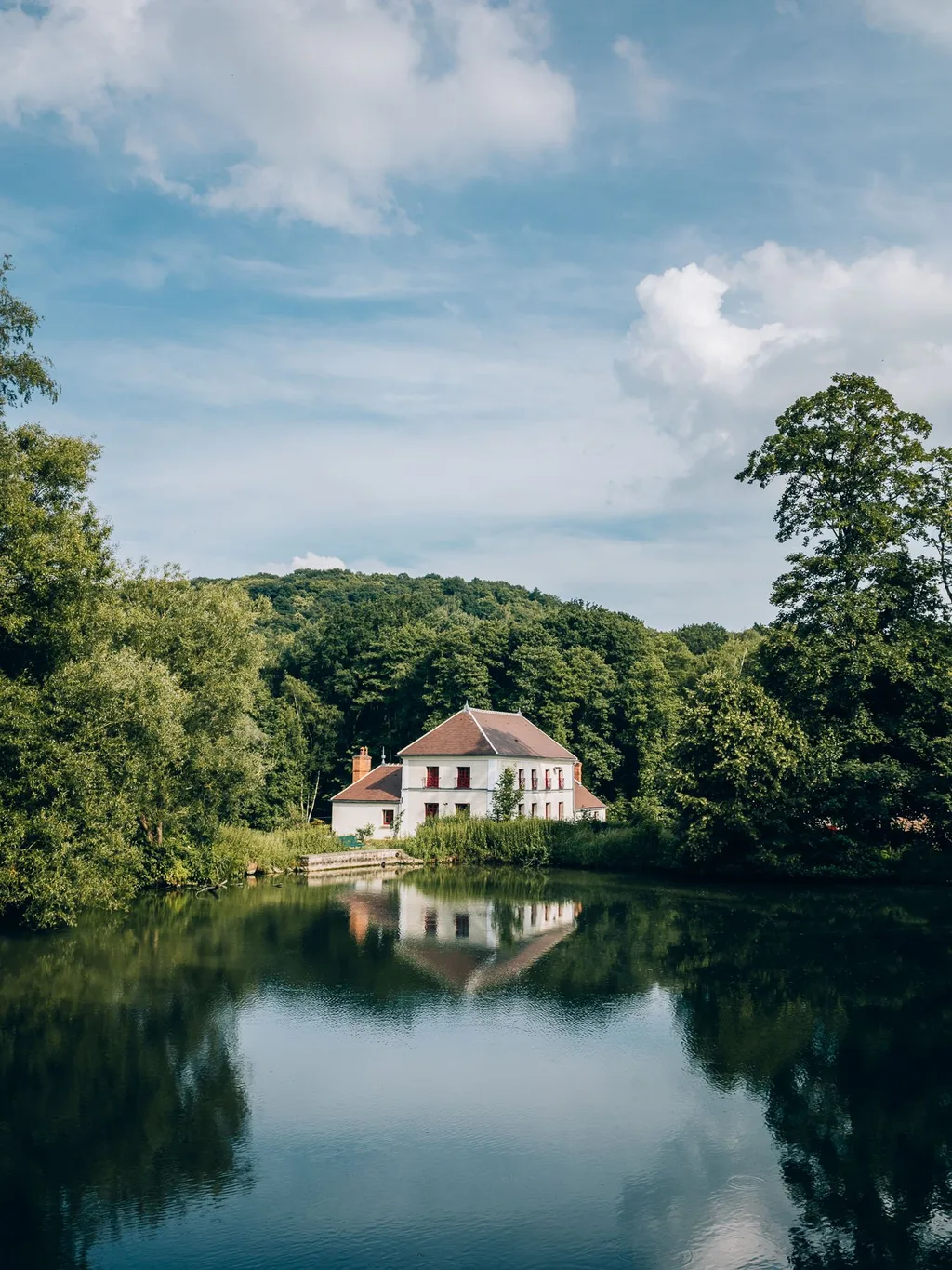 White manor house with red shutters mirrored in a still pond, Le Barn hotel