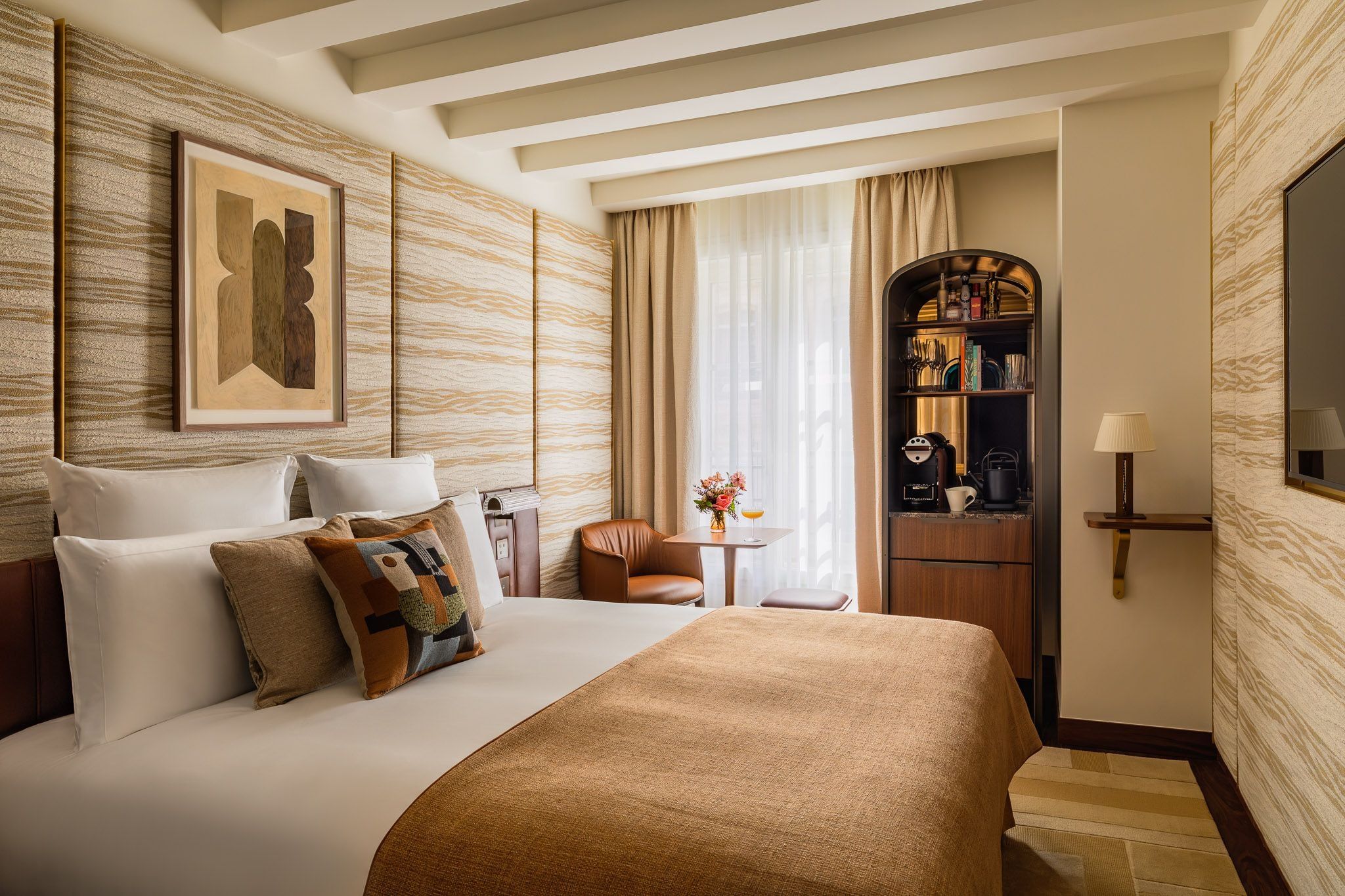 Bedroom with patterned beige walls, a rust-colored bedcover and white ceiling beams; a window-side table and chair sit beside an arched drinks cabinet.