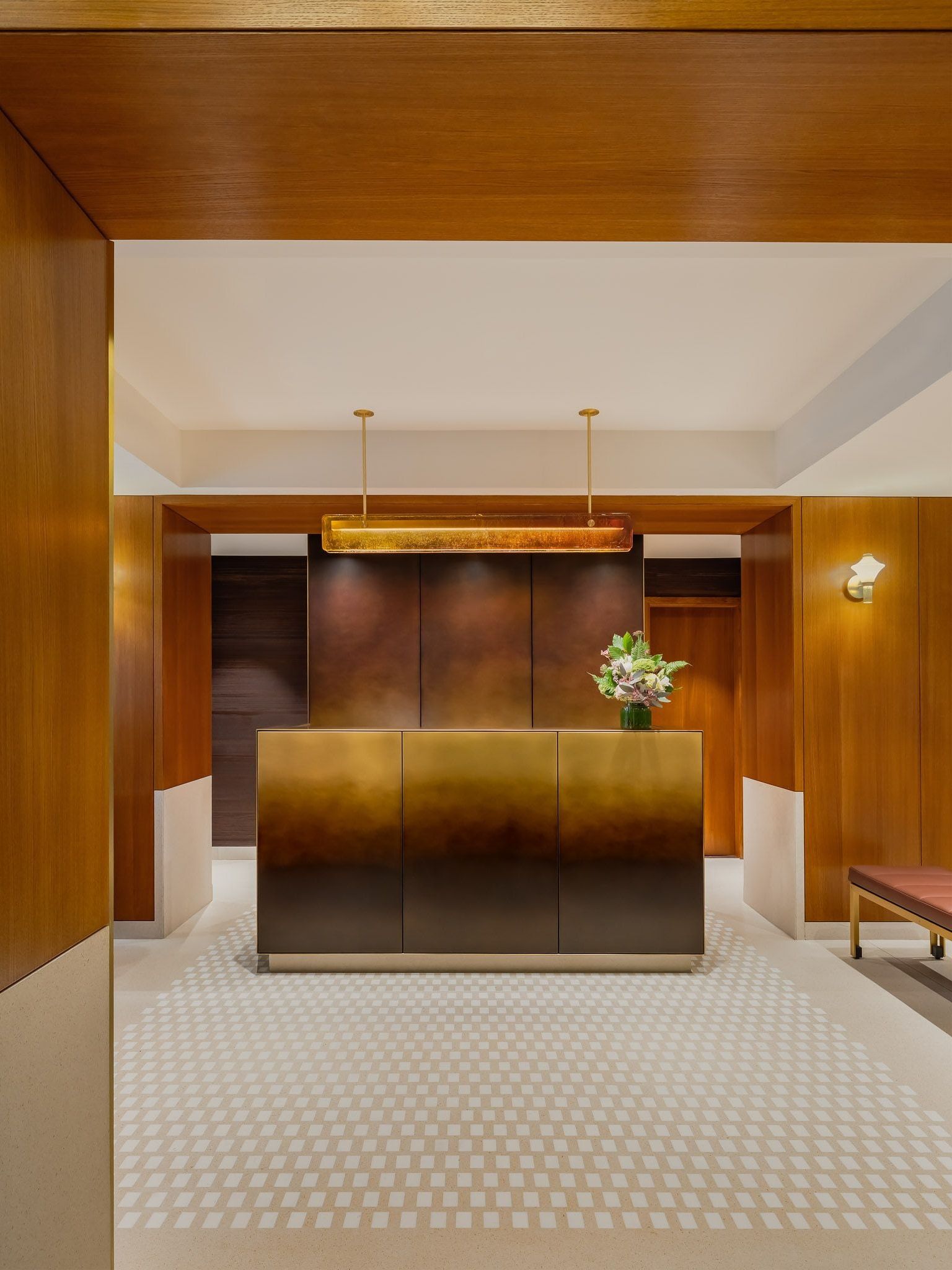 Reception desk with a gold-to-dark finish beneath a long pendant light, framed by wood paneling and a pale floor patterned with small white squares.