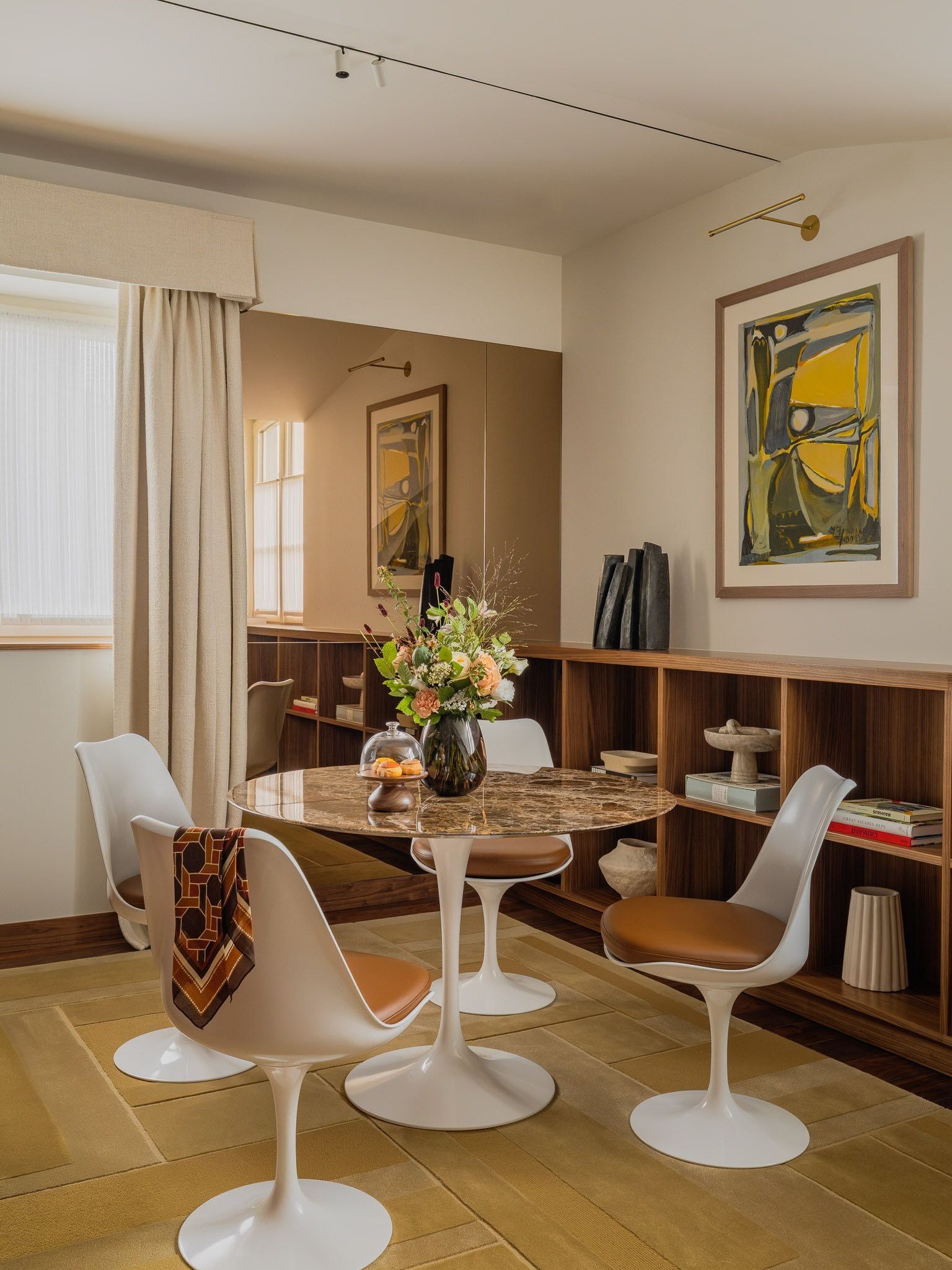 Round veined tabletop surrounded by four white pedestal chairs, beside low wooden shelving, a mirrored wall and yellow abstract artwork.