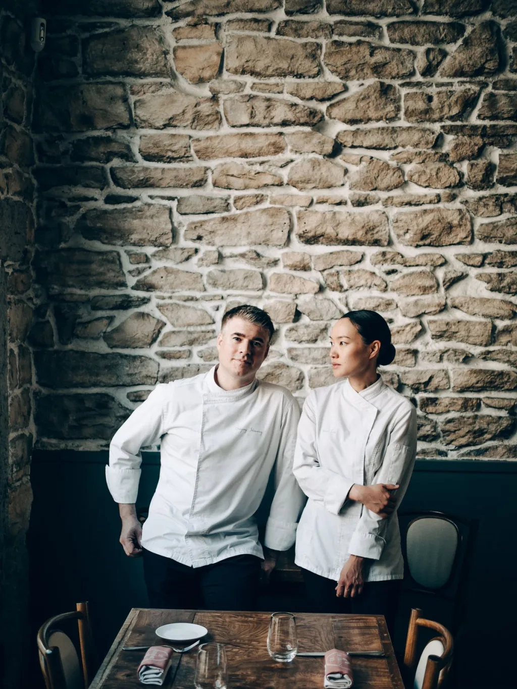 Two chefs in white jackets beside a dining table and exposed stone wall in Lyon