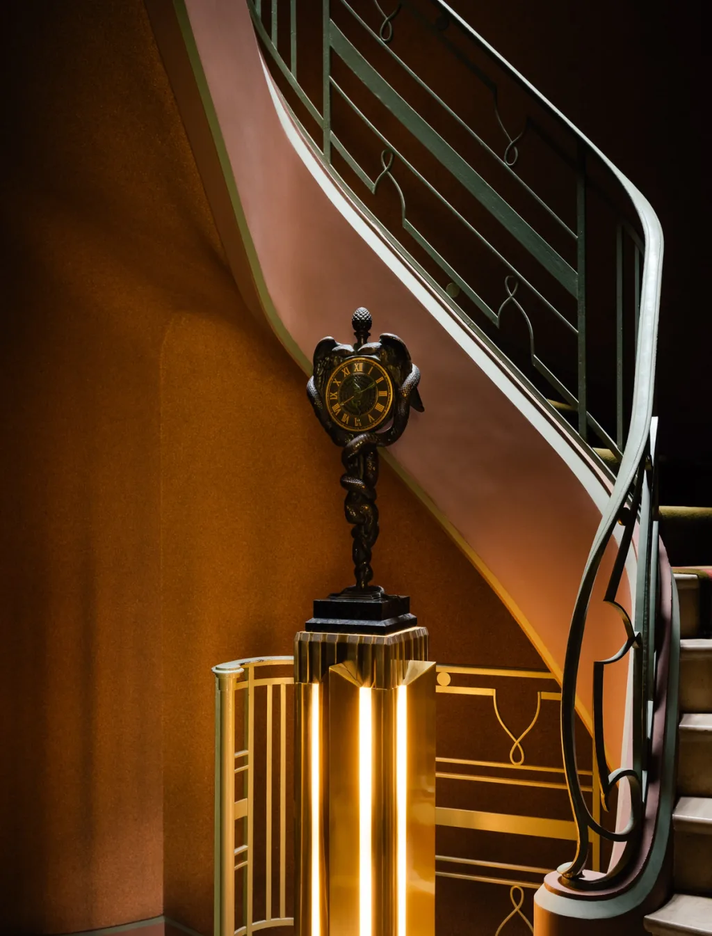 Ornate clock on a gold-toned pedestal beside a curving staircase, Felix Marcilhac series, Paris