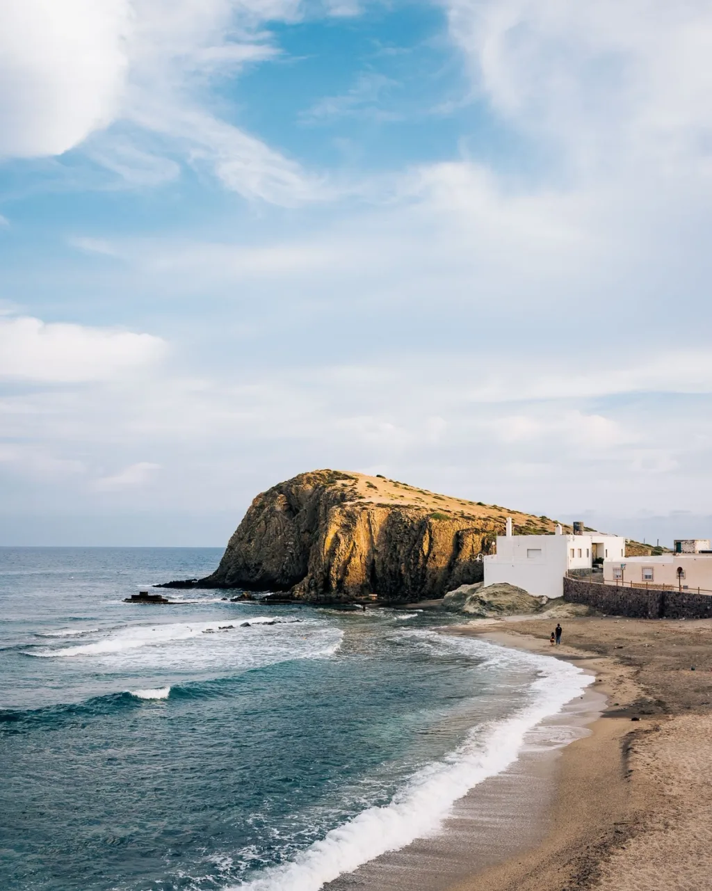 Waves wash a sandy beach beside white buildings and a rocky headland in Almeria