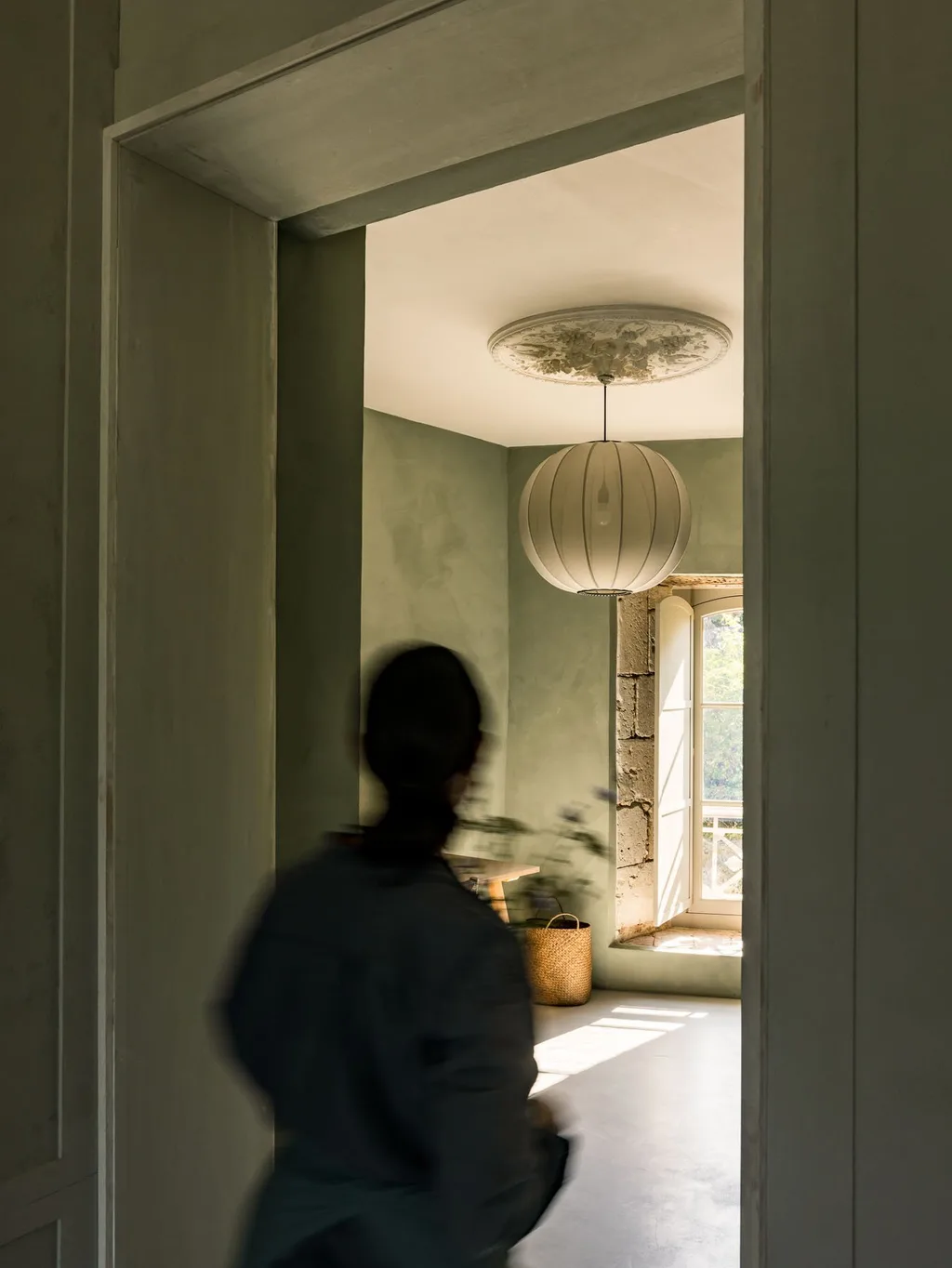 Christèle Ageorges walks past a round paper lantern in a green room, Manufacture Royale de Lectoure hotel