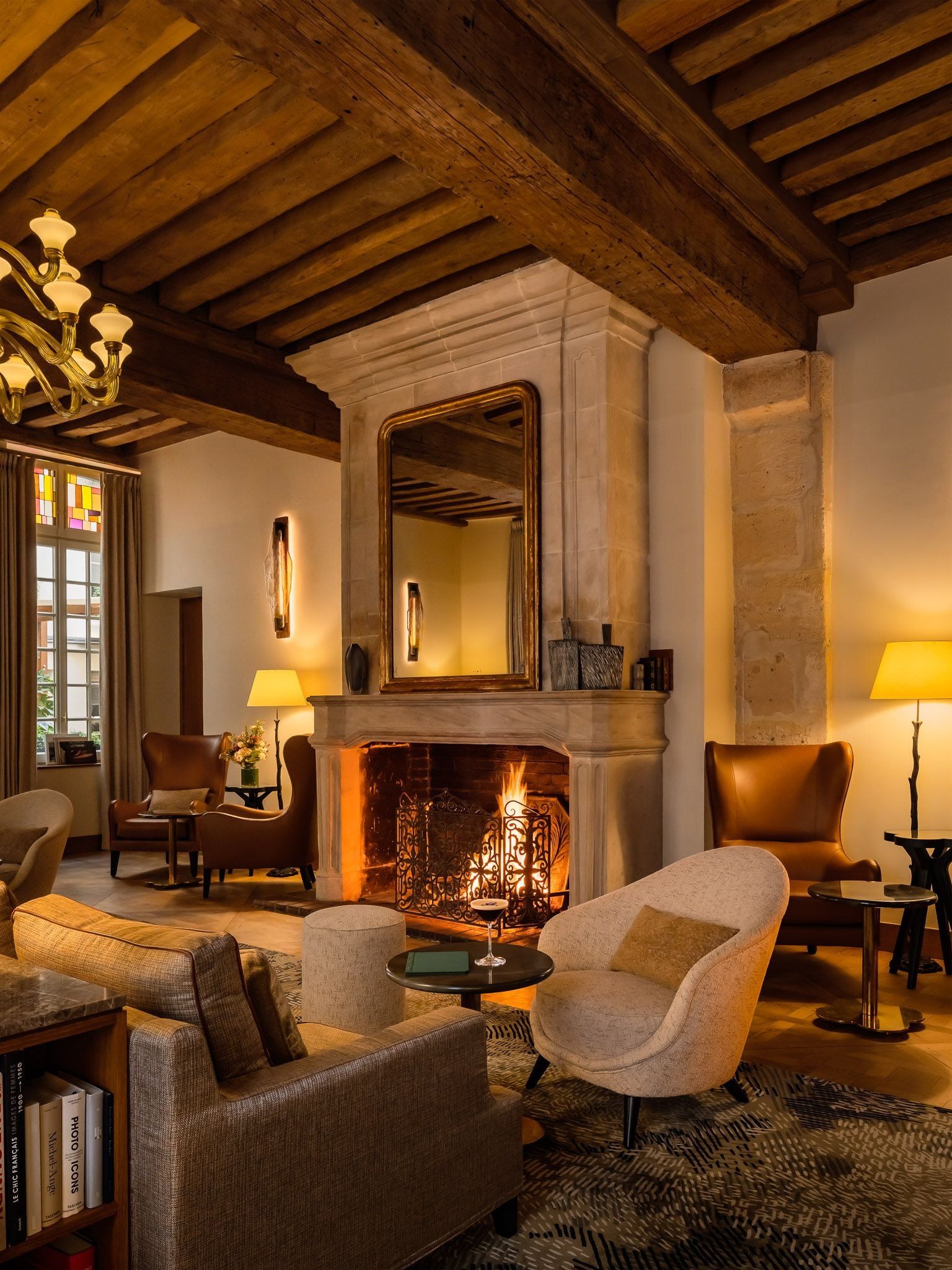 Lounge at Hôtel d’Aubusson, Paris, with exposed timber beams, armchairs around a lit stone fireplace and a tall gilt-framed mirror above the mantel.