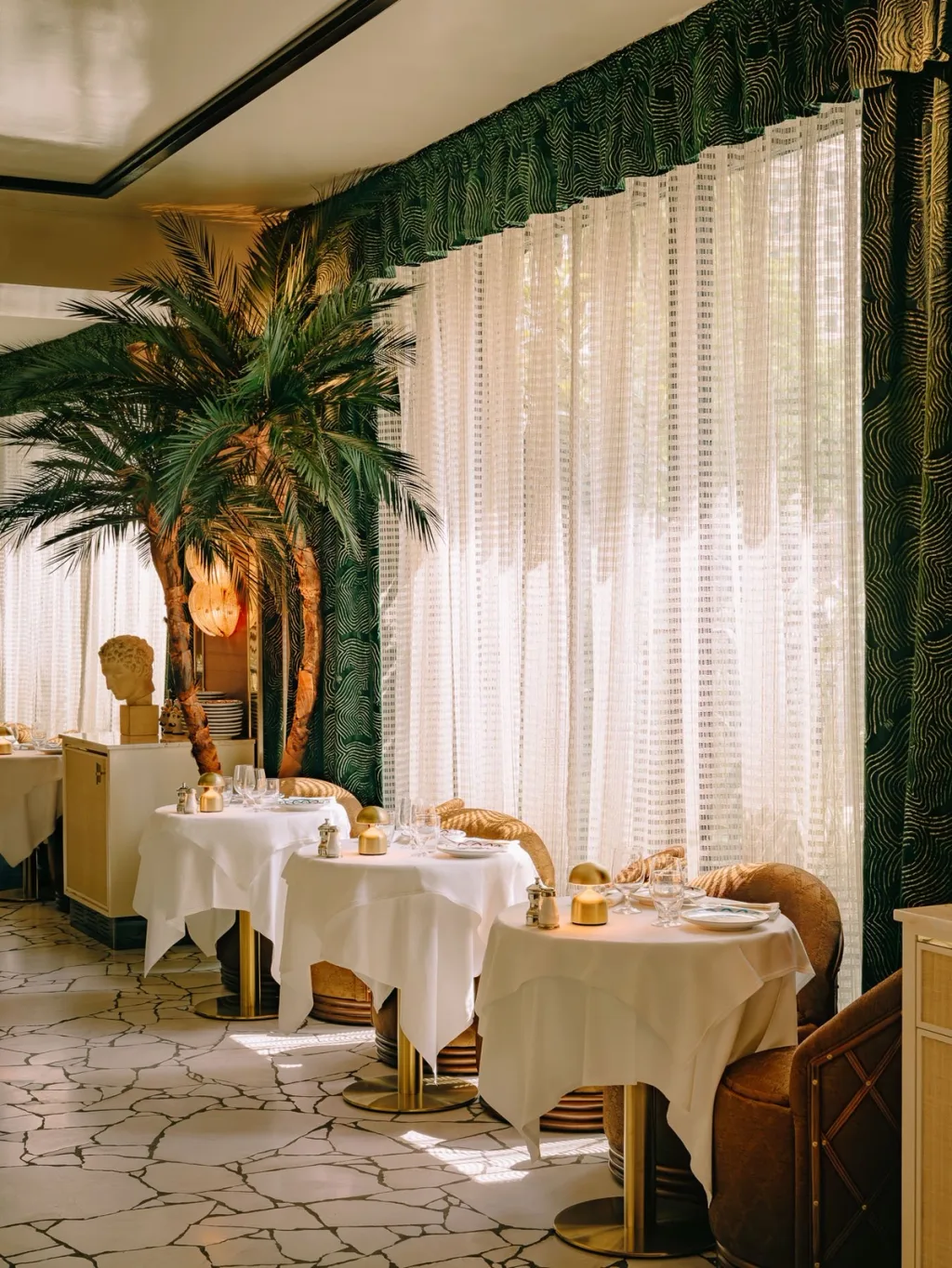 White-clothed dining tables beside tall sheer curtains and a palm tree at Big Mamma, UK