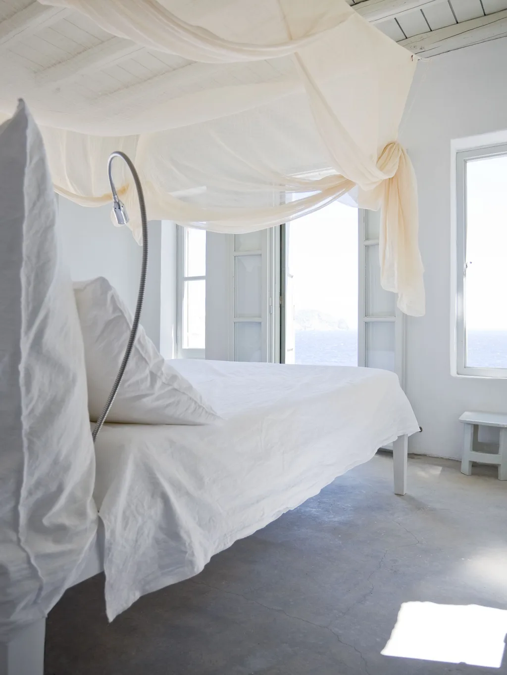 White bed beneath loosely draped fabric beside sea-facing windows, Paola Navone, Serifos