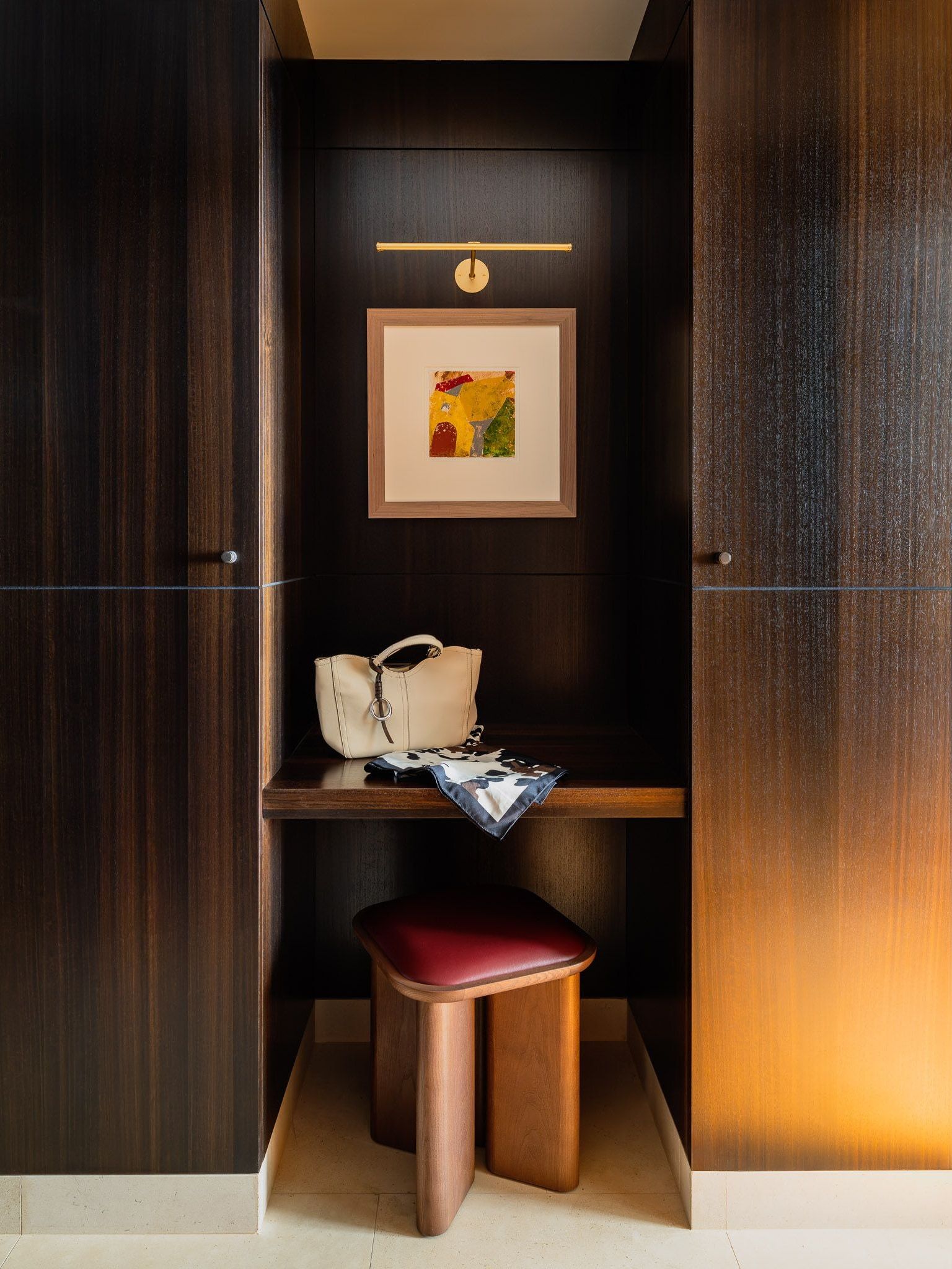 Dark wood alcove between cupboards, with a handbag and scarf on a shelf, a burgundy-seated stool below and framed abstract art above.