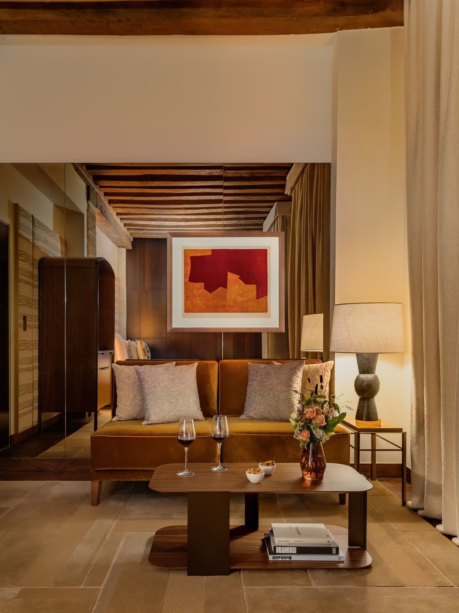Ochre sofa behind a low table with wine glasses and flowers, framed by a mirrored wall, exposed ceiling beams and red abstract artwork.