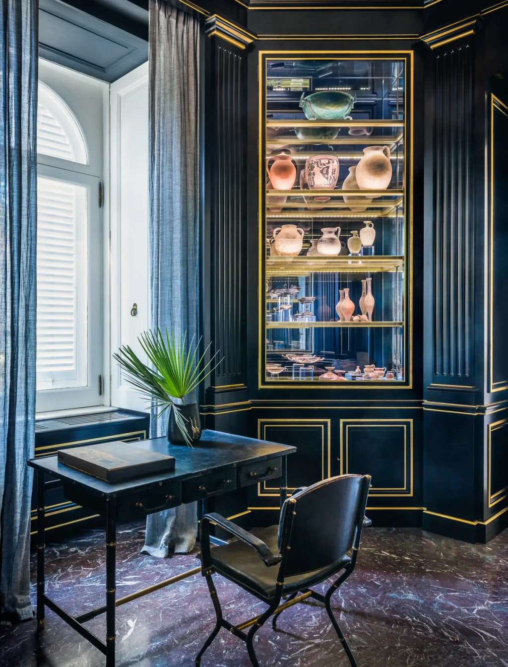 Small desk beside a lit display of vessels in a dark blue room at Villa Astor
