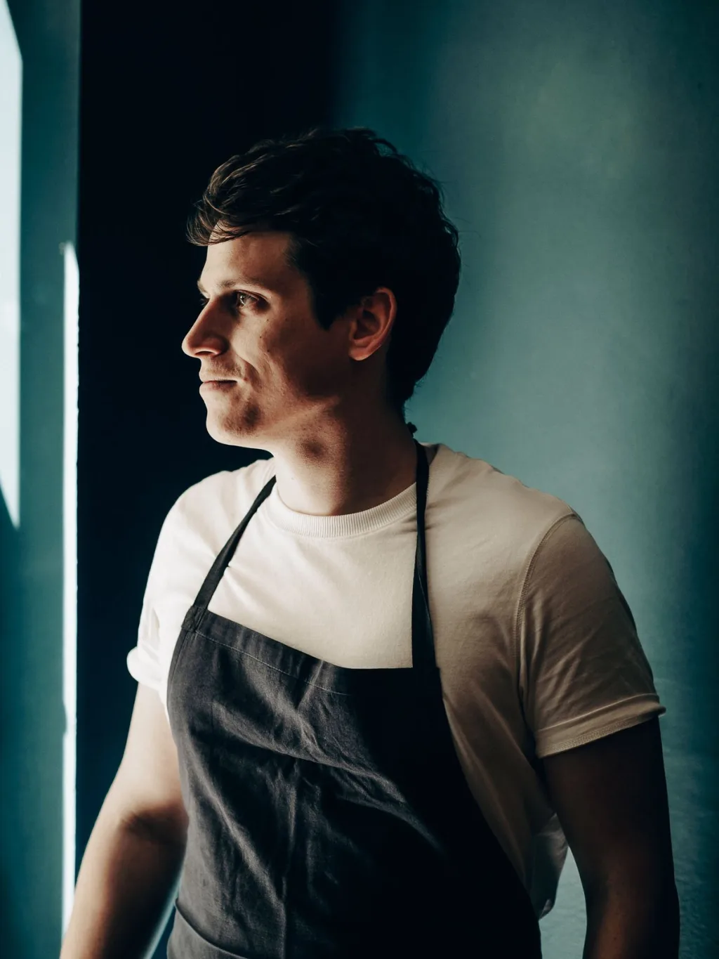 Man in a dark apron standing in side light against a blue-green wall in Lyon
