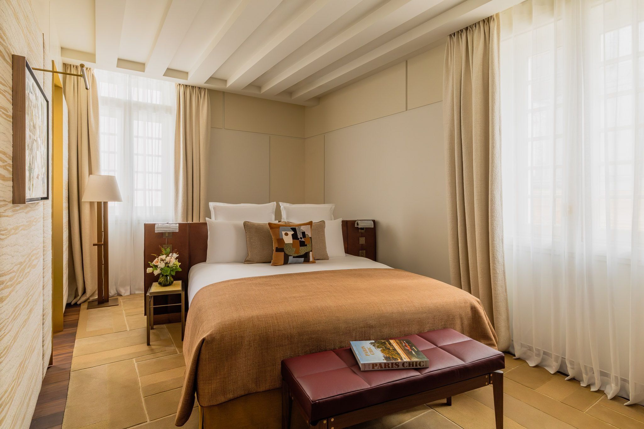 Bedroom at Hôtel d’Aubusson, Paris, with a rust-colored bedcover, burgundy bench and tall curtained windows beneath white ceiling beams.