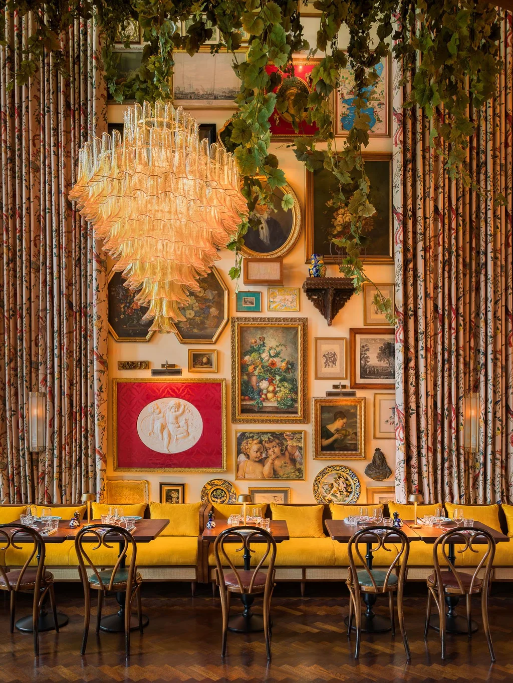 Tall chandelier, hanging foliage and an art-filled wall above a yellow banquette at Big Mamma, UK