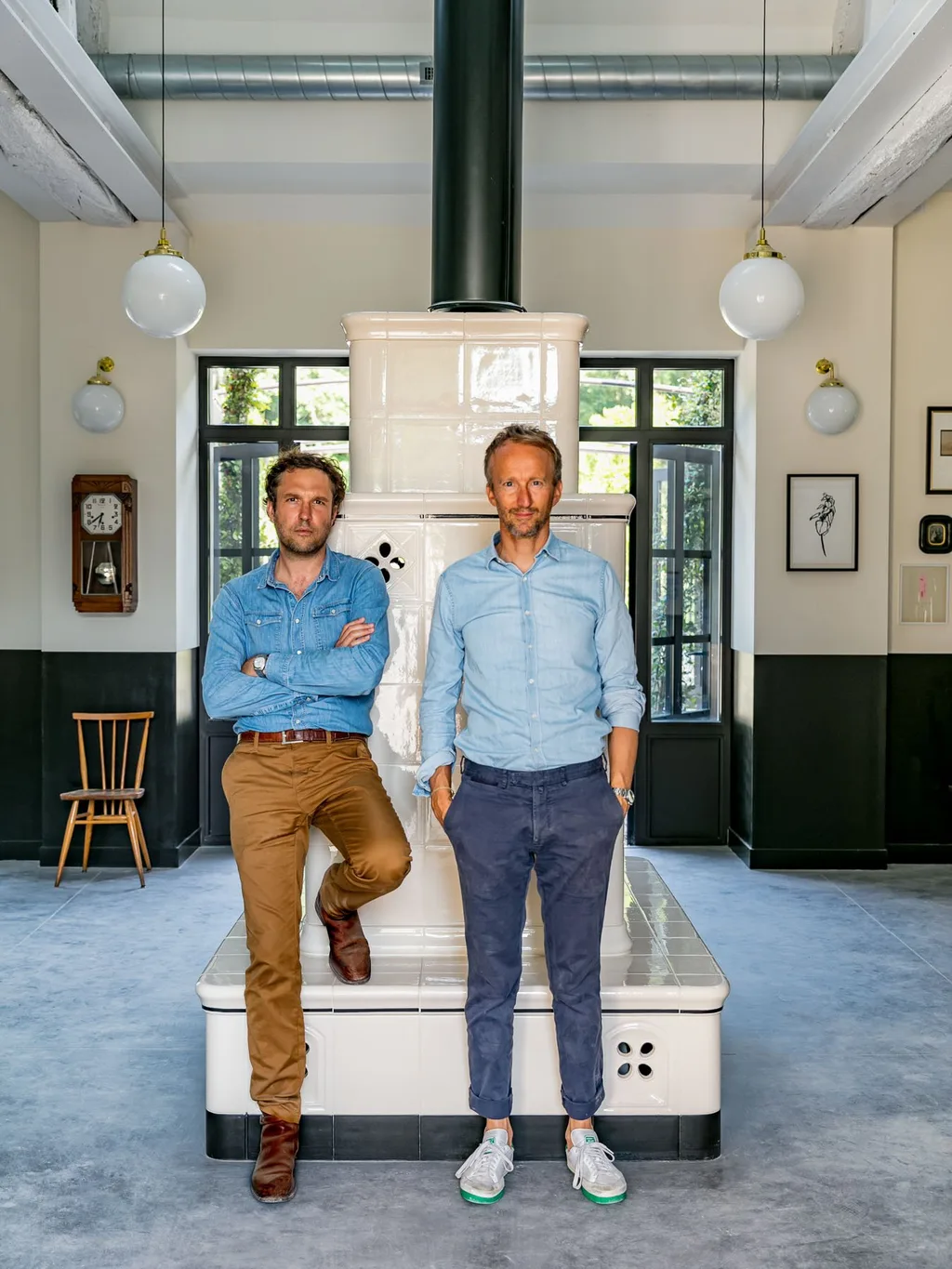 Two men lean against a tiled wood stove in an industrial lounge, Le Barn hotel