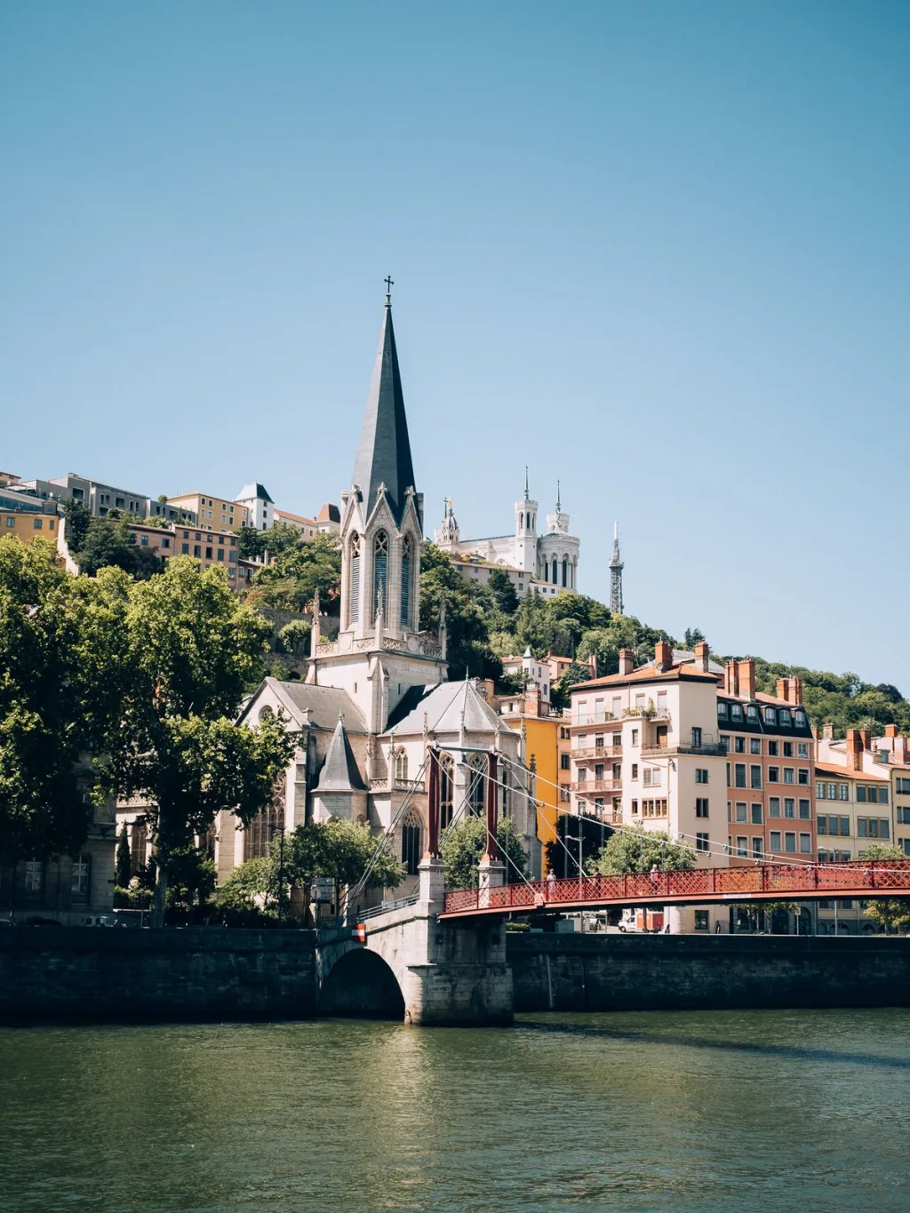 Church spire and hillside buildings beside a red pedestrian bridge over the river in Lyon