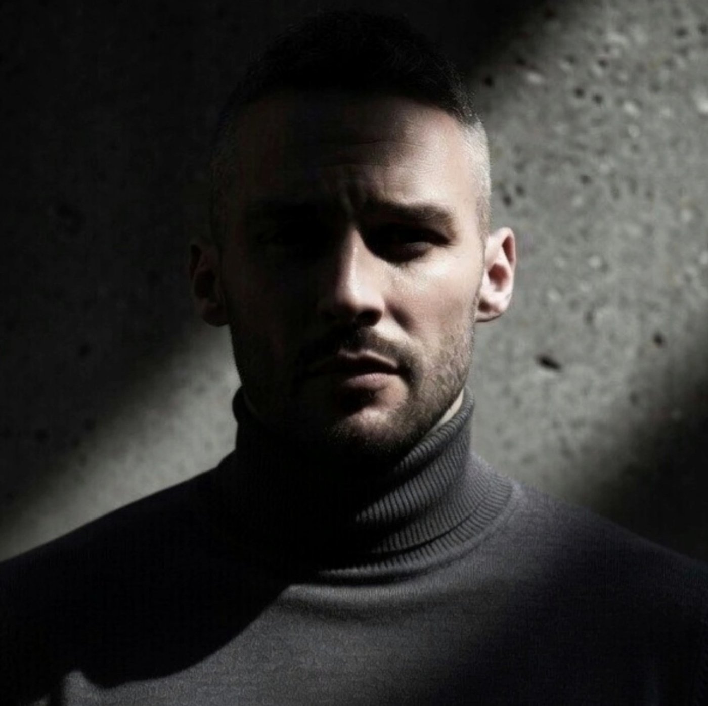 Phil G. Pavlov in a charcoal turtleneck, dramatically lit against a textured wall