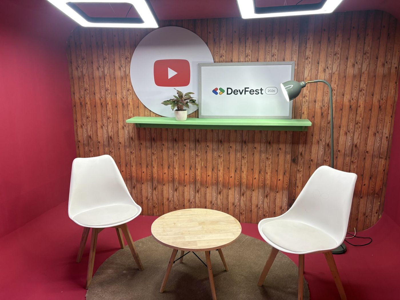 A lounge corner at the Google office set up for DevFest — YouTube and DevFest branding on a wood-panelled wall, a plant, and two chairs around a small table.