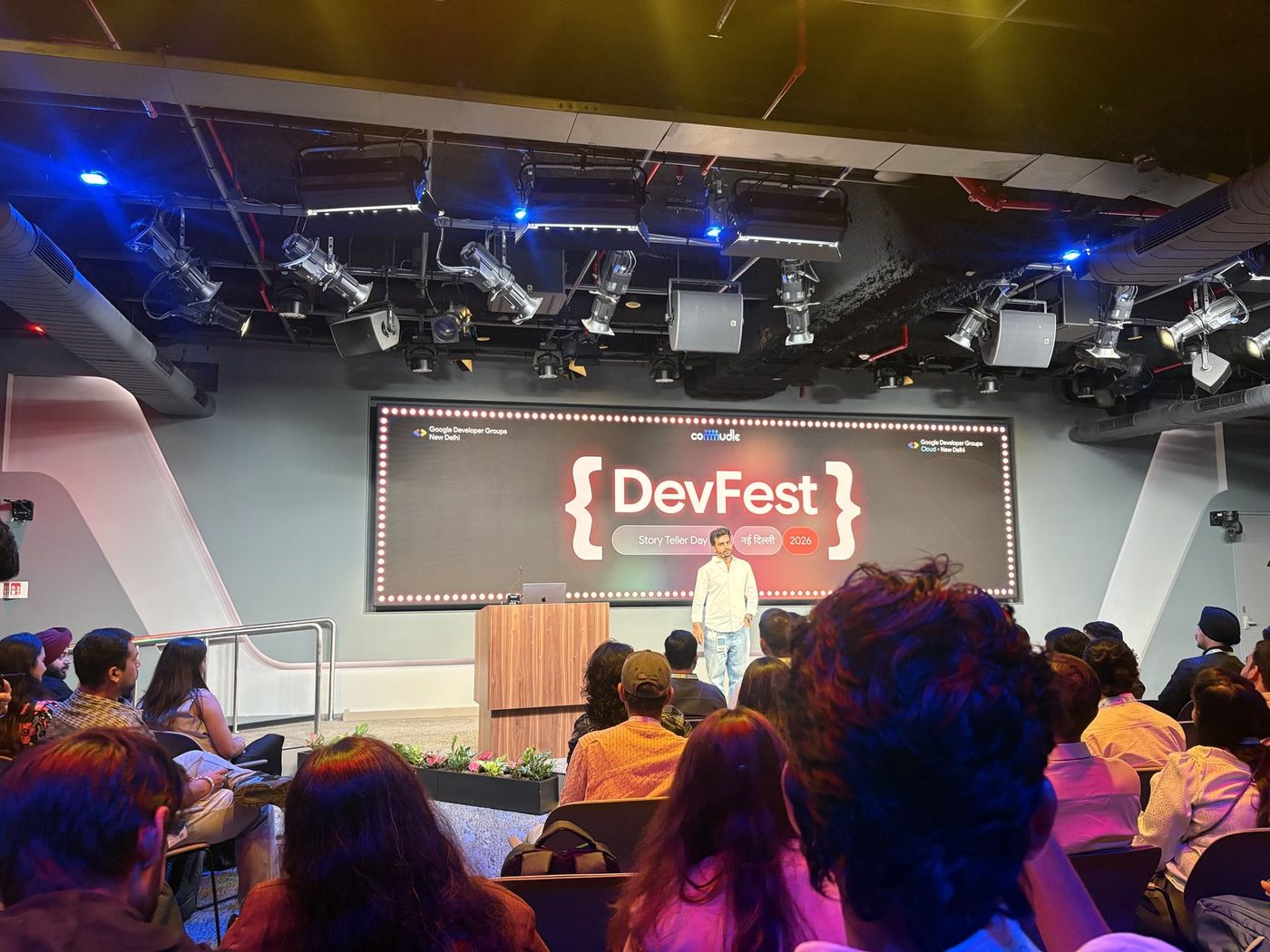 Wide shot of the DevFest hall — a seated audience facing a speaker on a lit stage under a large DevFest banner.