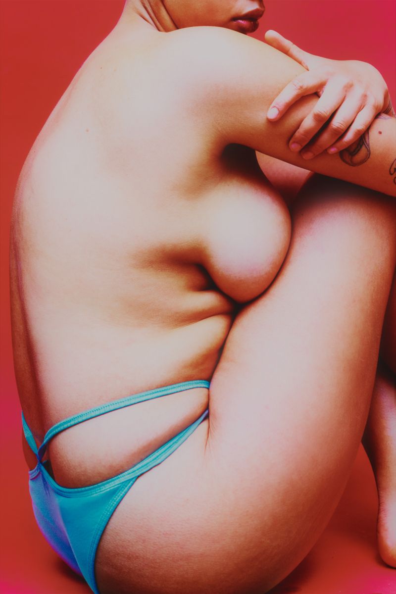Model sitting on red backdrop bugging body wearing blue pants