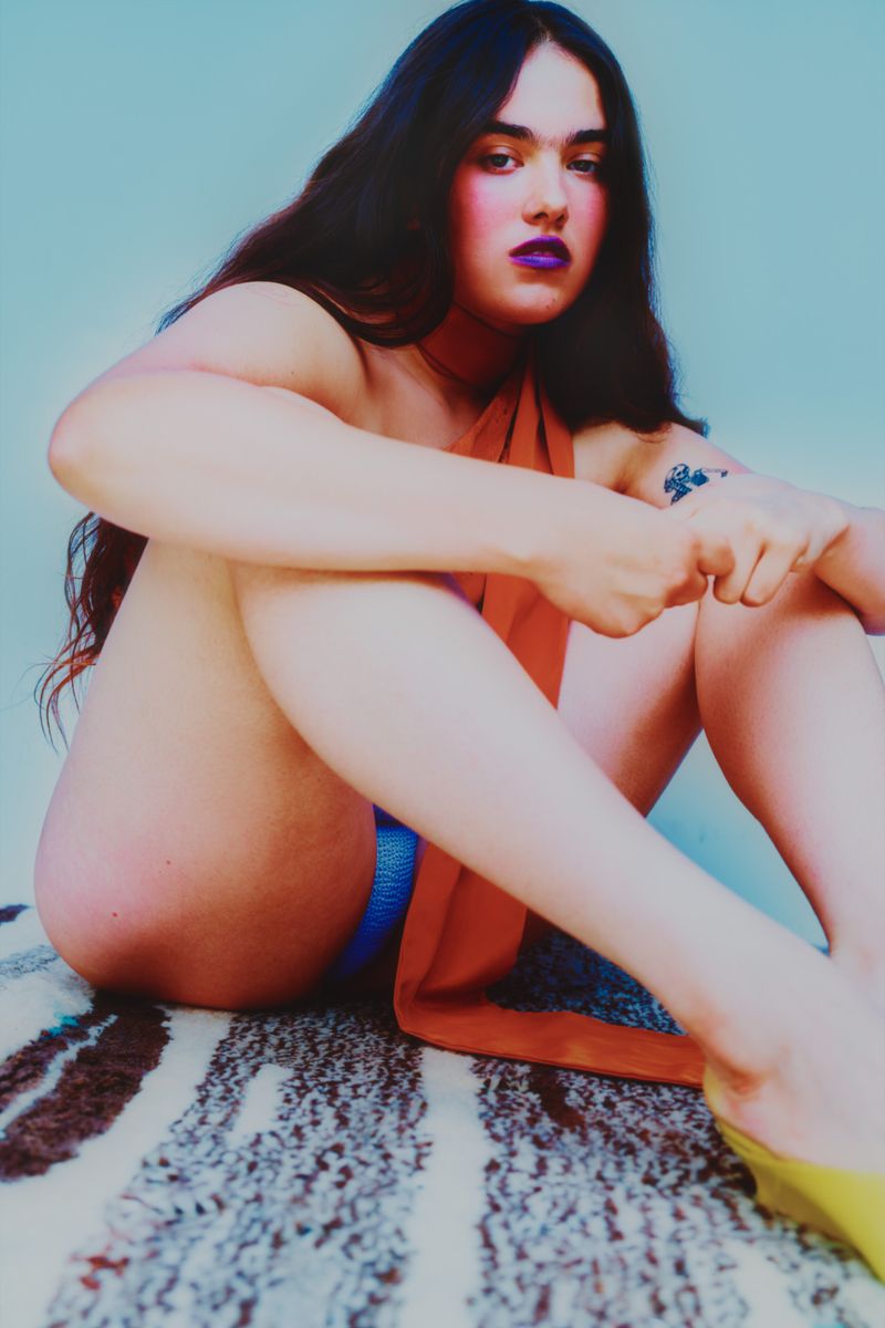 model sitting on a neutral striped rug wearing orange top, and yellow shoes