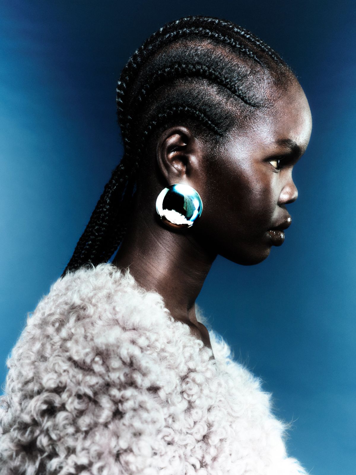Model on blue background wih braids in her hair and a fluffy beinge coat, big silver earrings