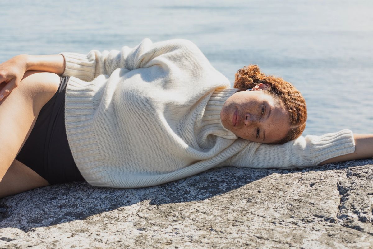 women lies across a rock near the sea wearing a jumper
