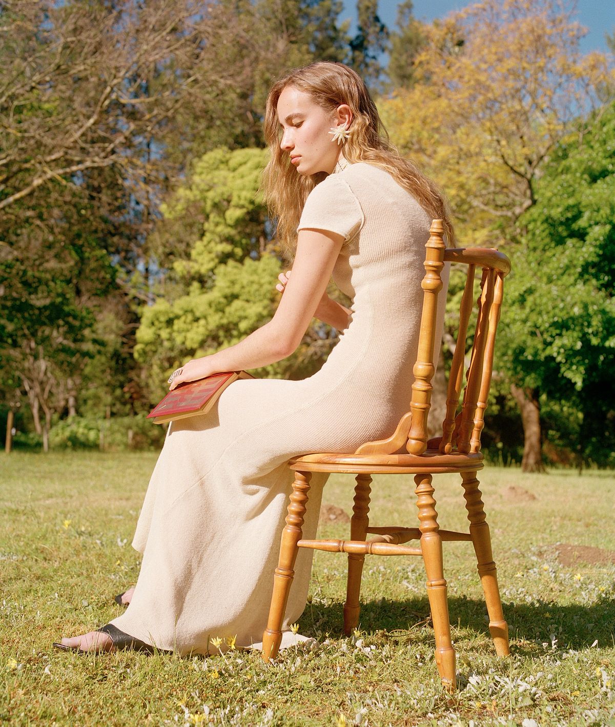 A model wearing a cream dress sitting on a chair on the grass holding a red book