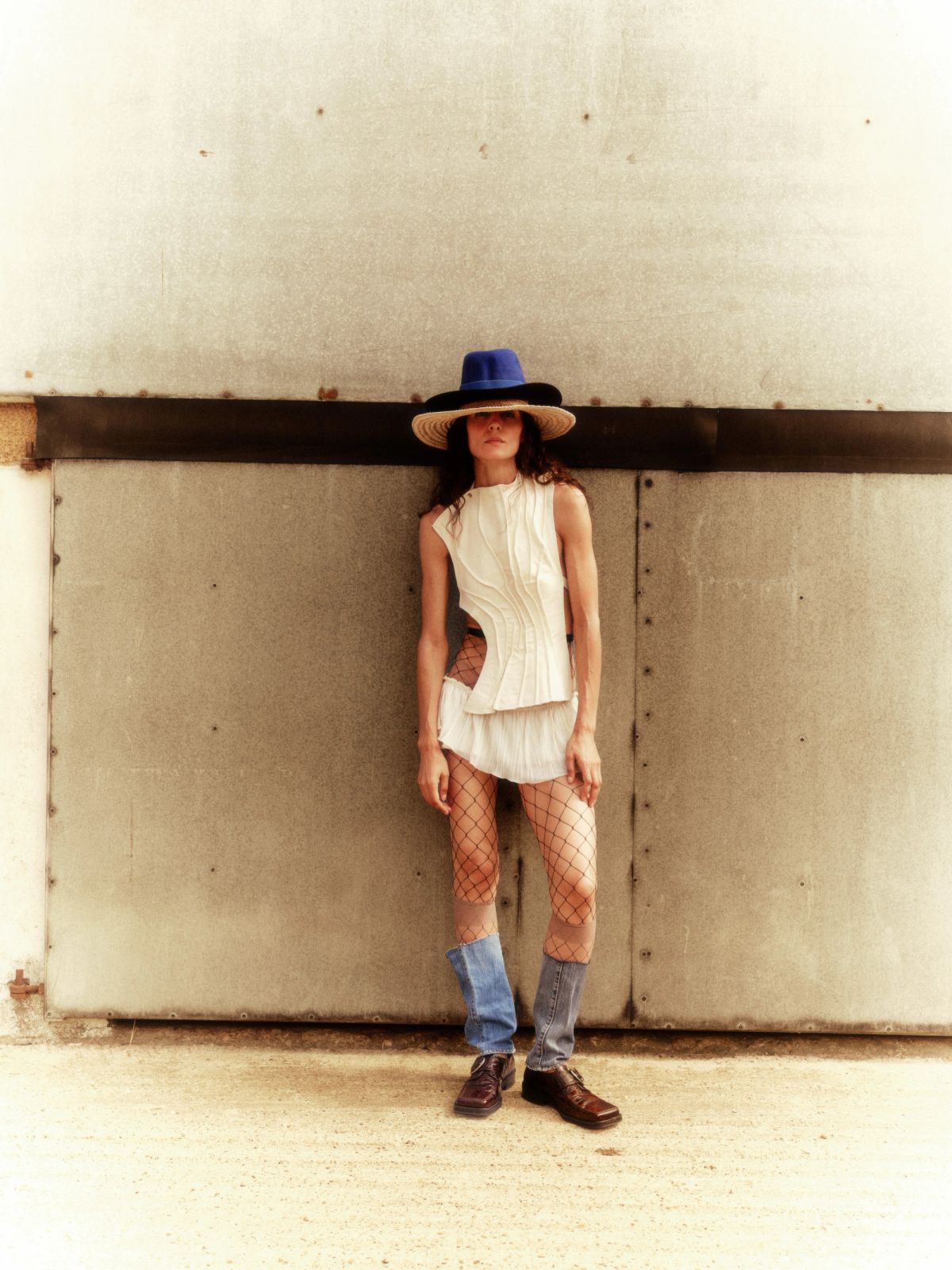 Model standing on the side of a farm barn wearing two hats and cream clothing