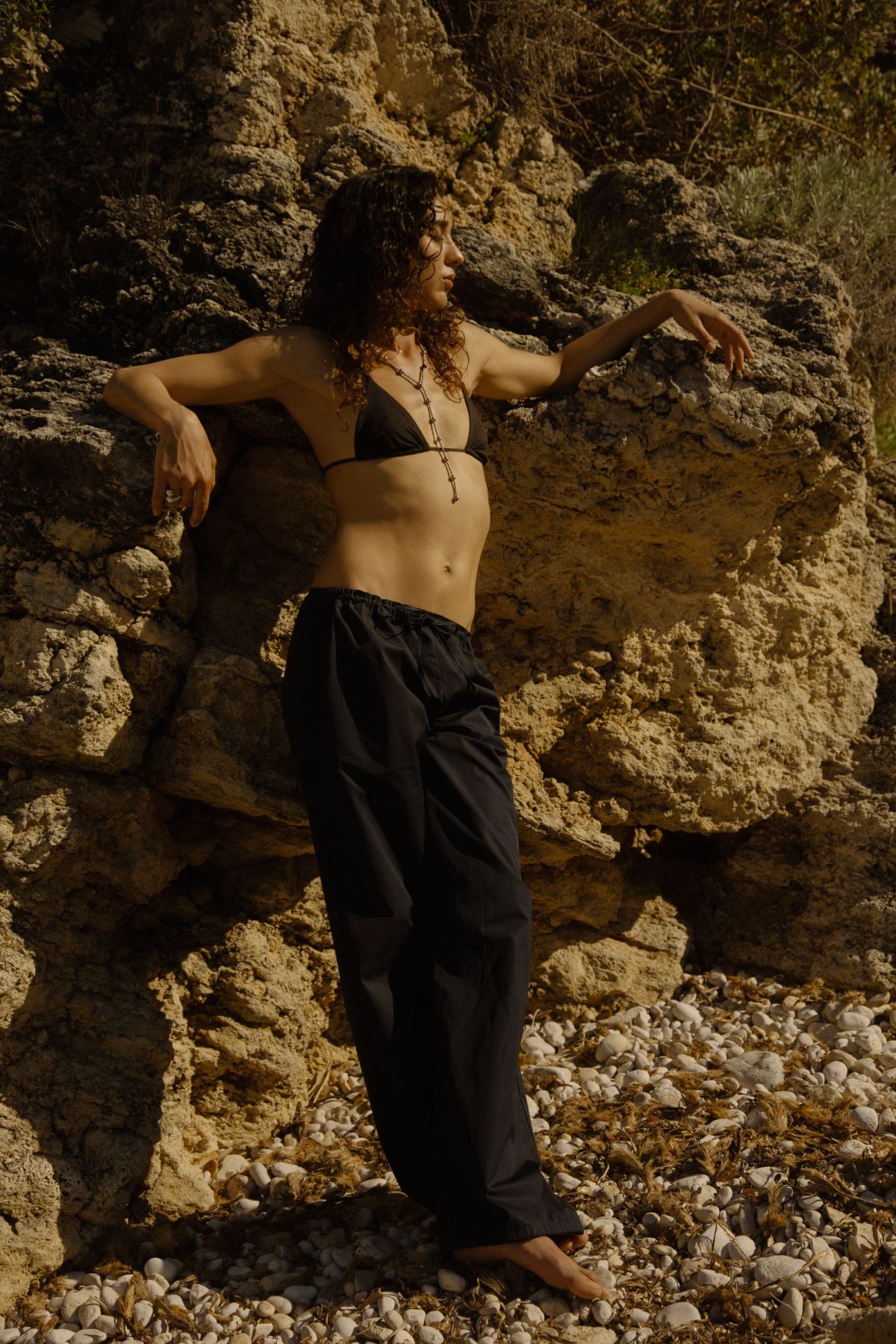 Woman standing at rocks in black bikini and black trousers