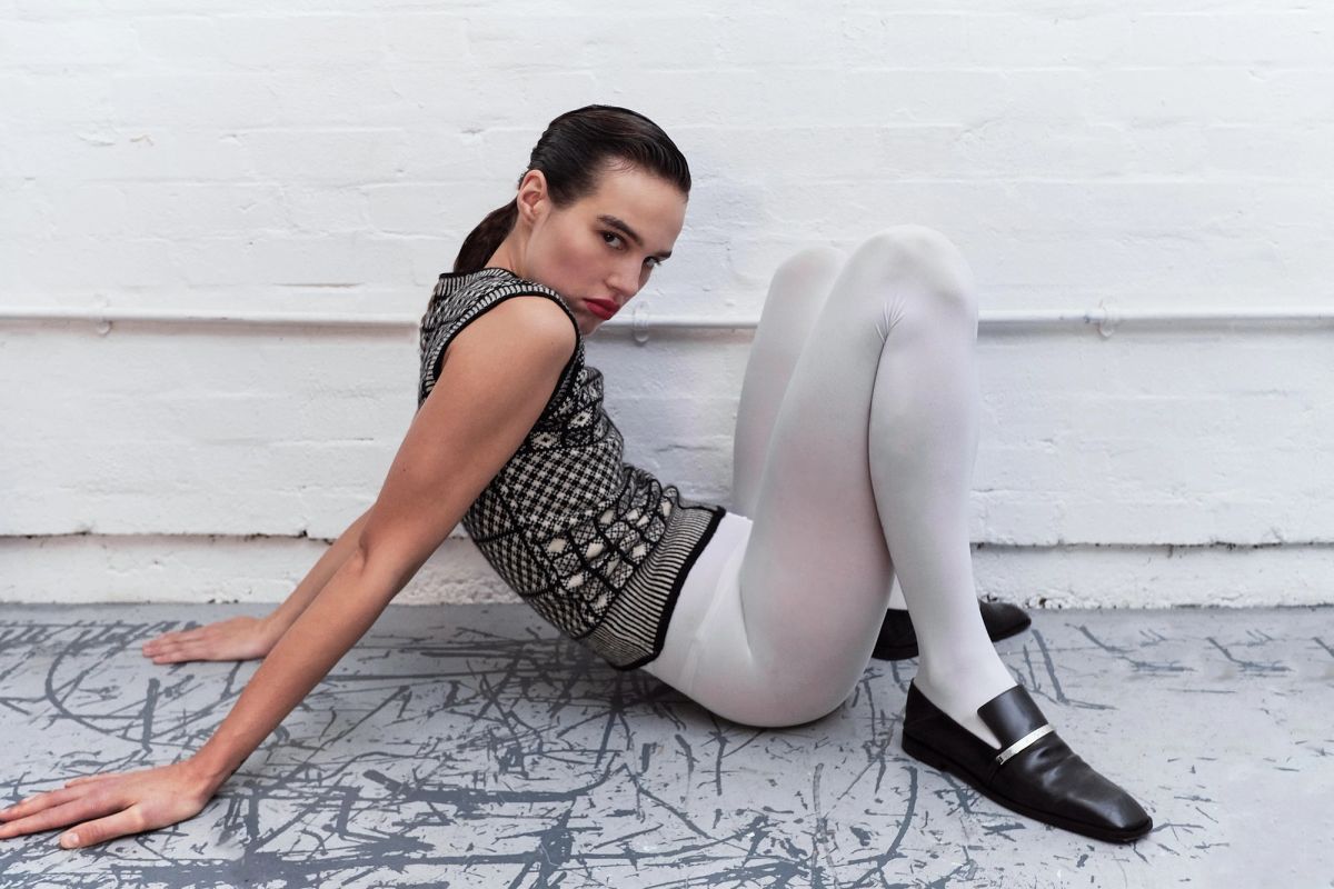model sitting on a scratched grey floor wearing white tights and knitted vest