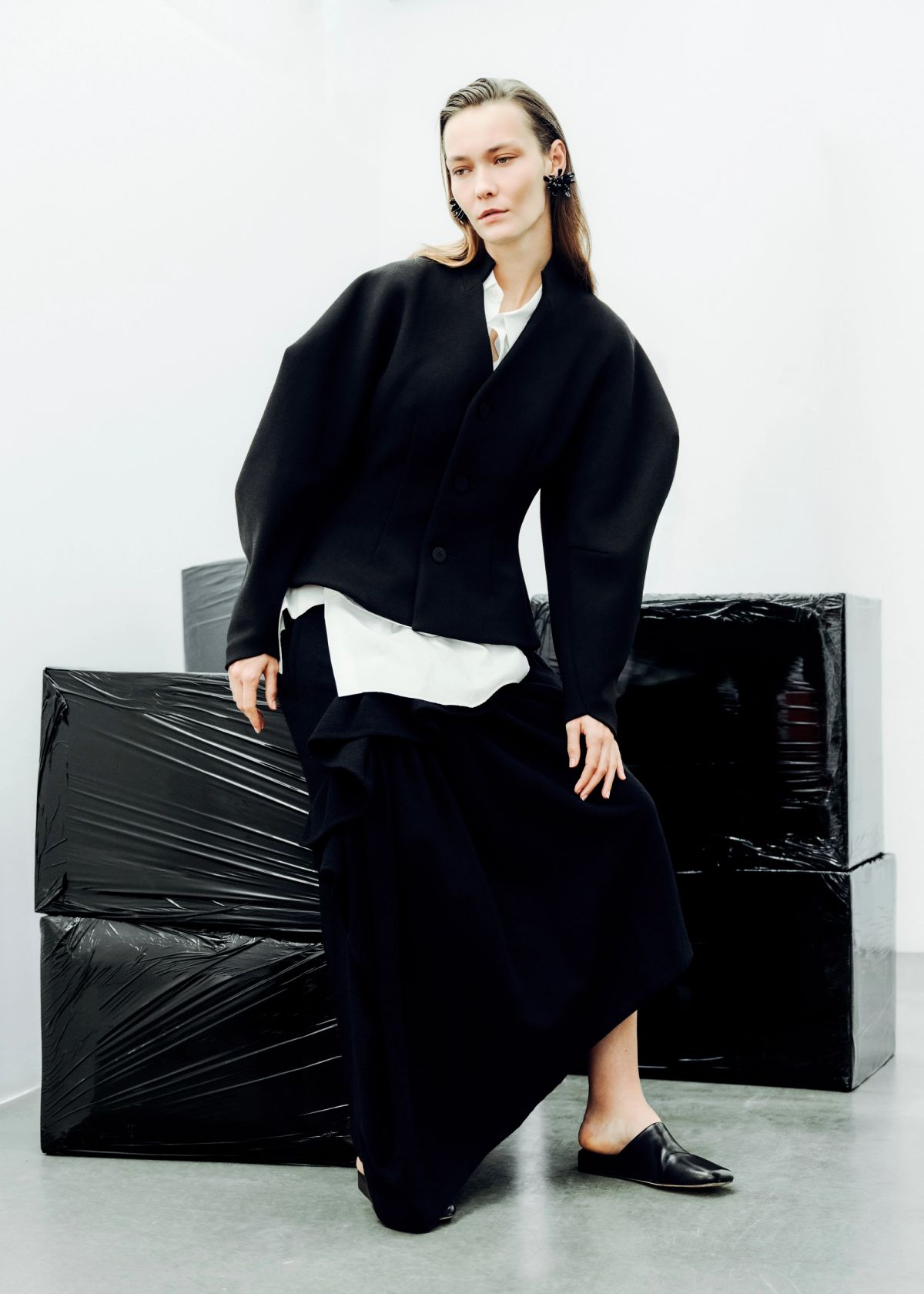 model wearing black skirt suit and white shirt with black wrapped plastic objects in the rear