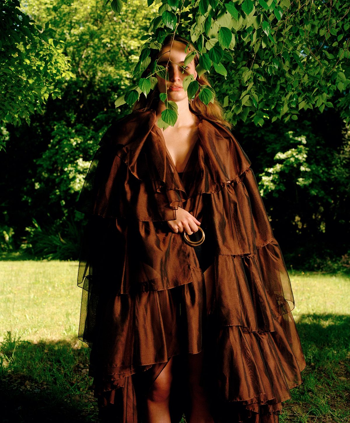 A model wearing a brown ruffled dress standing outside surrounded by green grass, a tree obscures her eye