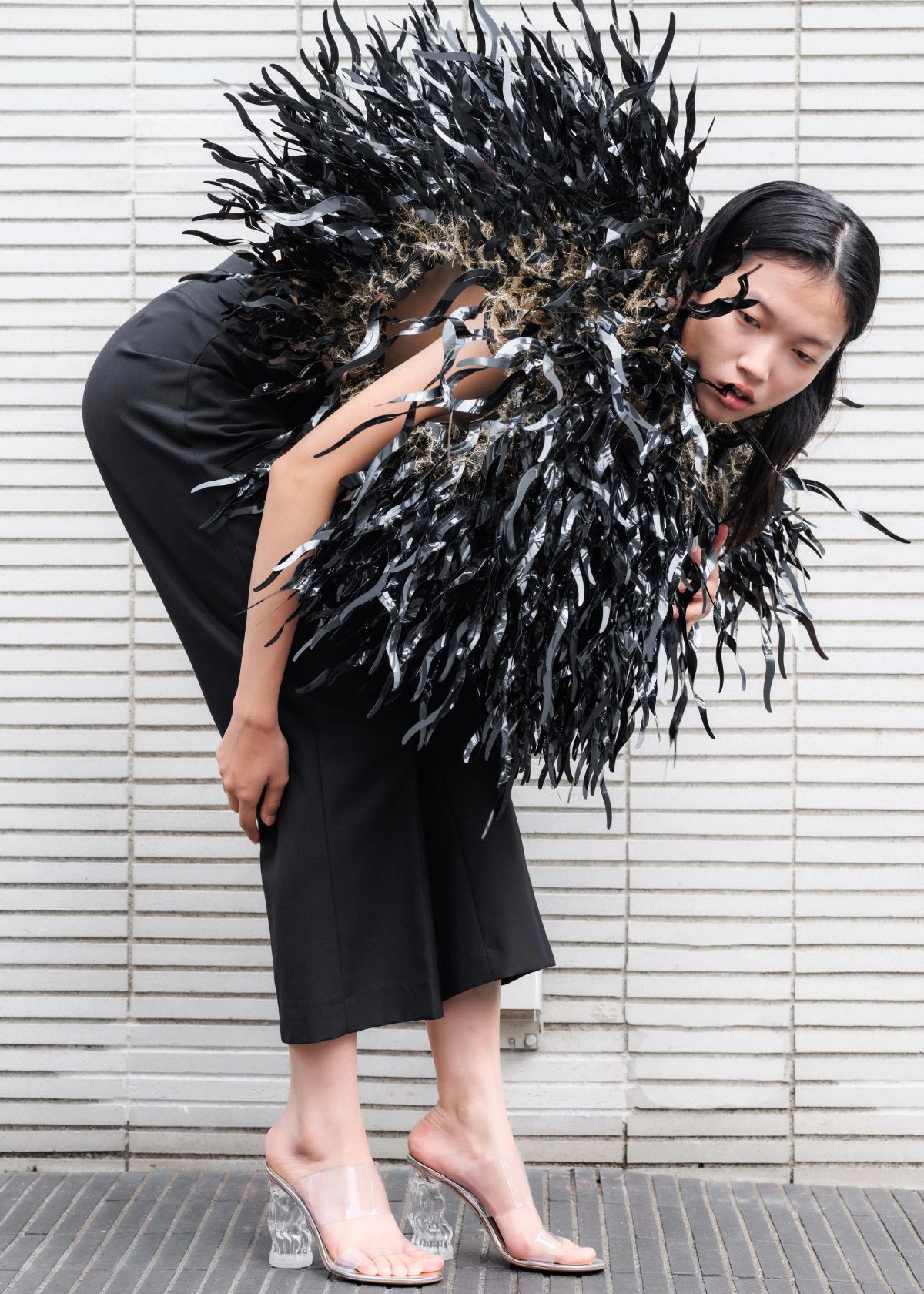 model wearing a black dress with a lot of texture bent over by a tiled white wall