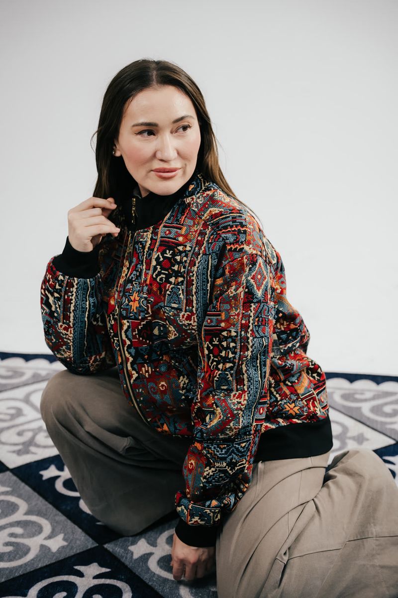 Banu Gaidar in a BAY bomber, seated on a rug with Kazakh ornament