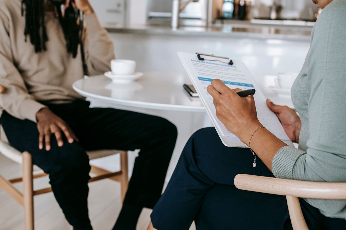 A caseworker's hands marking a printed screening checklist on a clipboard, a seated resident out of focus across the table.