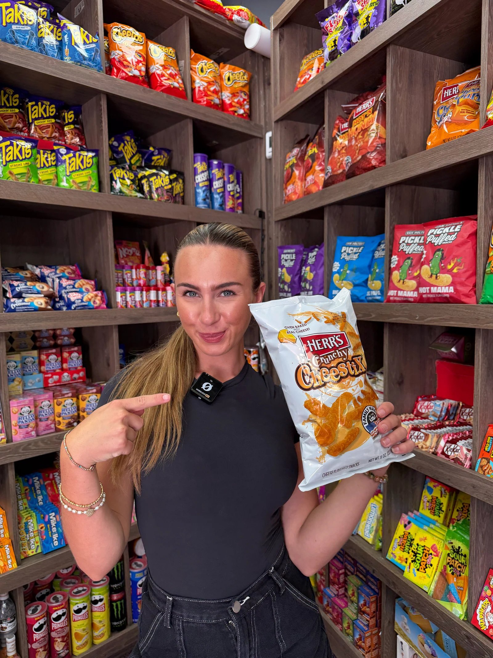 A Quirky Candy presenter reacting while holding a snack packet beside the shop shelves