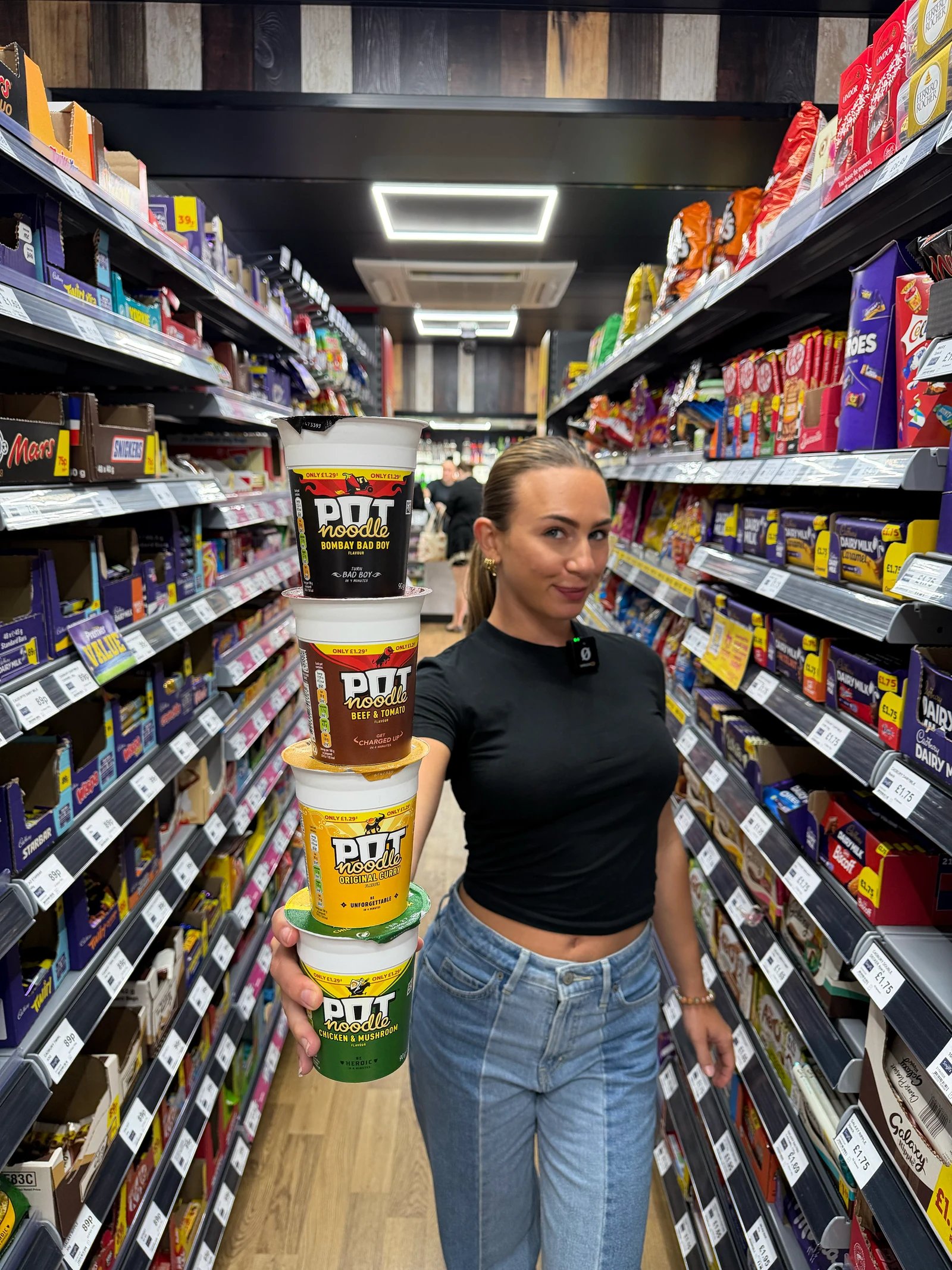 A presenter holding a stack of Pot Noodle tubs in a Singh's shop aisle