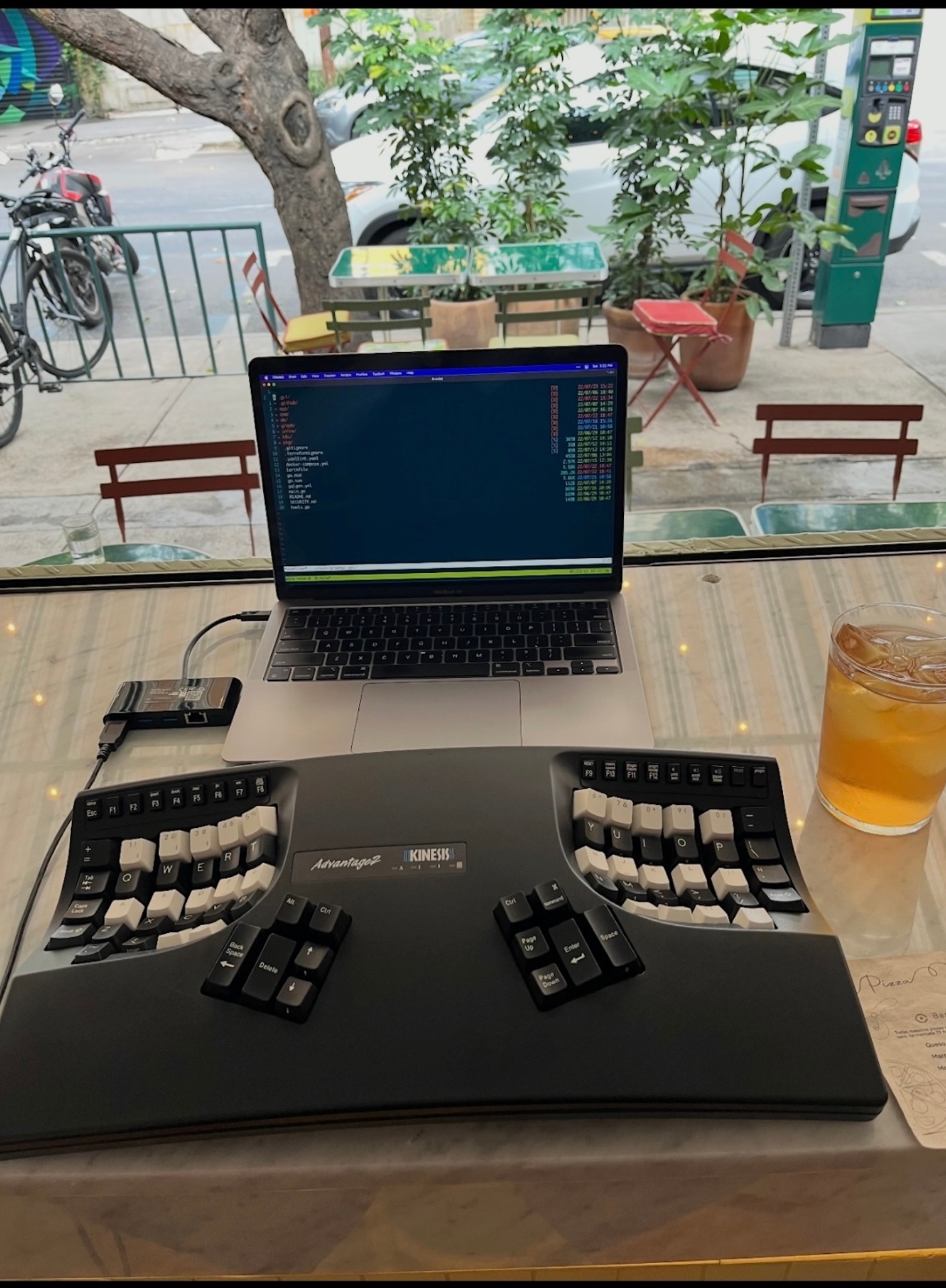 A compact café workspace setup with a laptop displaying a terminal interface in a tiling window manager, connected to a full-sized Kinesis Advantage2 ergonomic keyboard. The keyboard features concave key wells and thumb clusters for improved typing comfort, with black and white keycaps. The setup is placed on a marble tabletop next to a glass of iced tea and a small business card. Outside the window, urban street scenery is visible with parked bikes, potted plants, metal chairs, and a tree partially framing the view, creating a relaxed, mobile work environment.