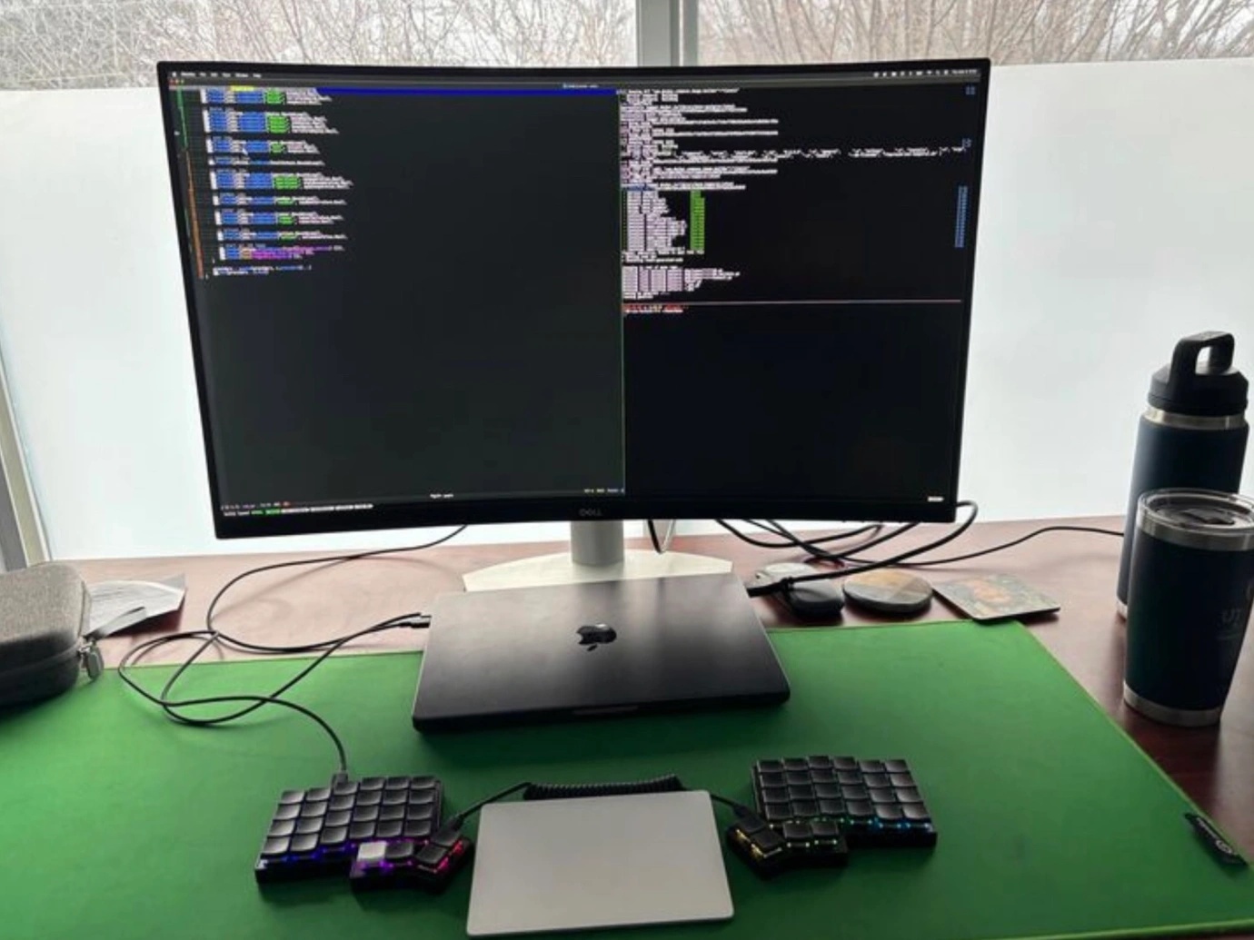 A clean coding workstation setup featuring a Dell ultrawide monitor displaying a split terminal interface with directory navigation on the left and command-line output on the right. Below the monitor is a closed MacBook connected to a split ortholinear mechanical keyboard with black keycaps and RGB backlighting, flanking a large Apple Magic Trackpad. The setup rests on a bright green desk mat. Additional desk items include a reusable water bottle, travel tumbler, and several coasters. Frosted windows and bare winter trees are visible in the background, suggesting a calm, focused remote work environment.