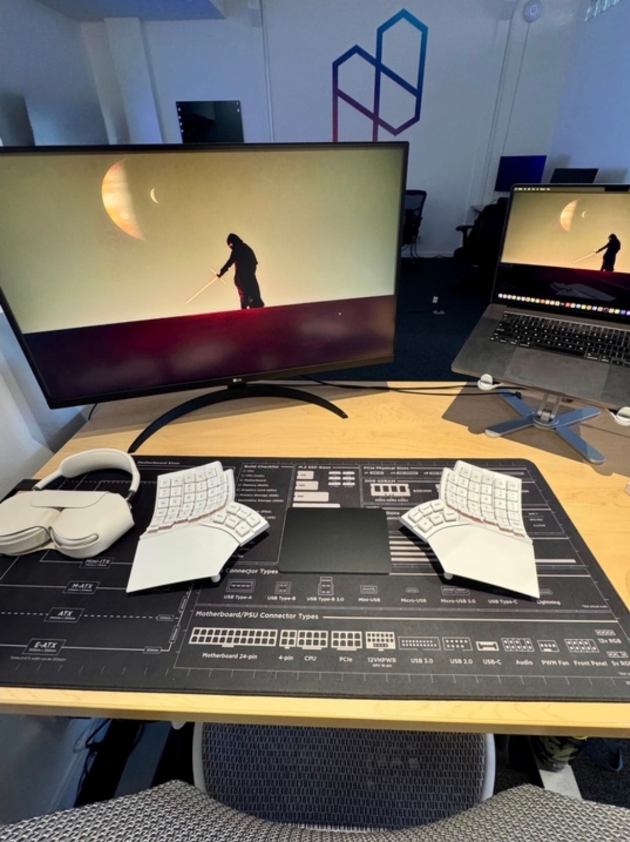 A modern office desk setup featuring a white split mechanical keyboard with curved key wells and a central black trackpad on top of a large black desk mat printed with motherboard and PSU connector diagrams. To the left is a pair of white over-ear headphones resting on the desk. The setup includes a MacBook on a stand connected to a large LG monitor, both displaying a minimalist sci-fi wallpaper of a figure with a lightsaber standing against a yellow sky with two moons. In the background, a wall features a geometric Nuon logo in gradient blue and pink lines, suggesting the workspace is part of a tech or developer office.