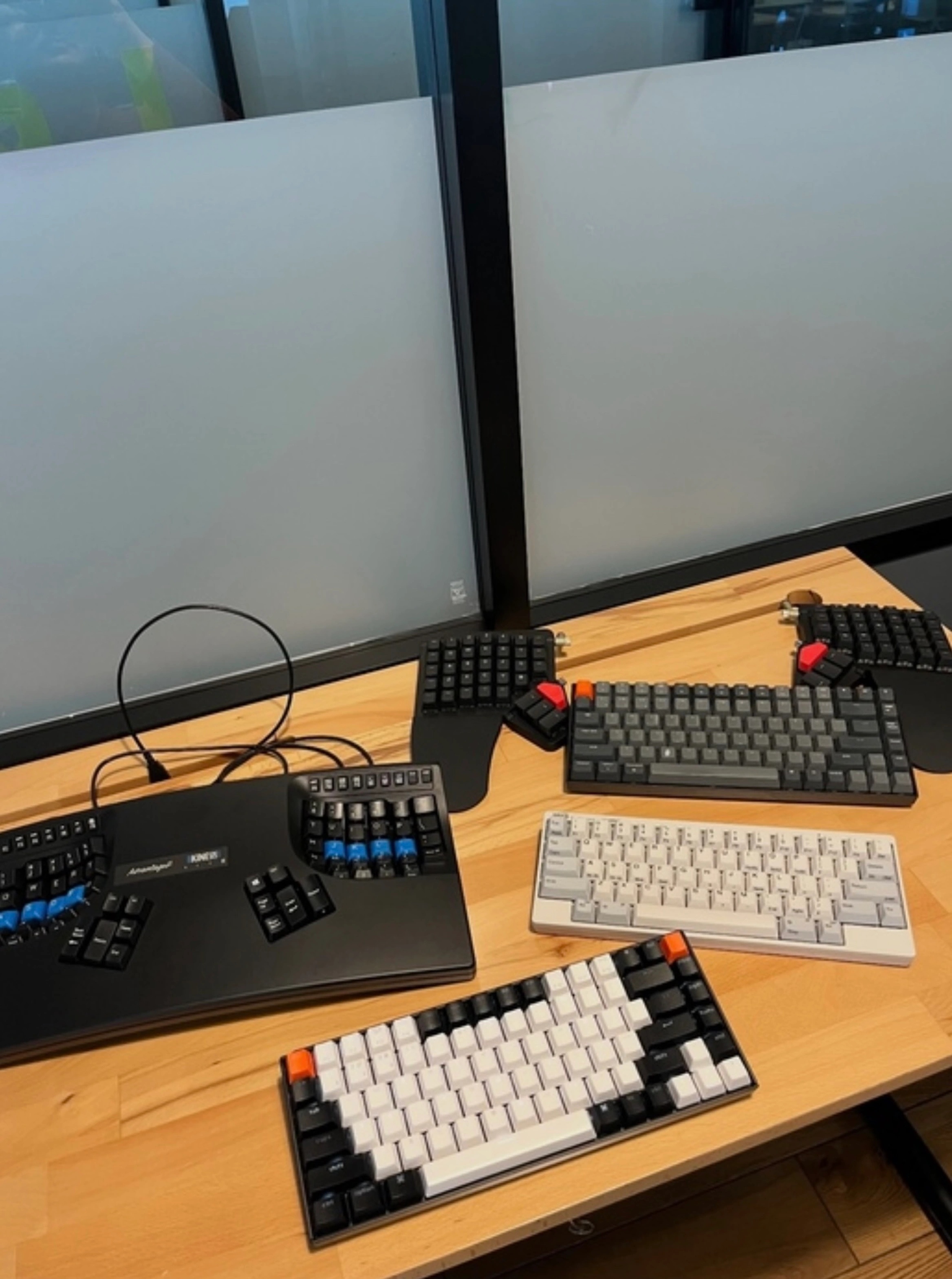 A collection of five mechanical keyboards arranged on a light wooden desk in an office setting with frosted glass dividers. Featured keyboards include a Kinesis Advantage2 ergonomic keyboard with blue-tinted key switches in the thumb cluster, a compact gray split keyboard with thumb clusters and black keycaps, and three 60–65% custom mechanical keyboards in various color schemes—gray-on-gray, white-and-gray, and white-and-black with orange accents. The setup highlights a variety of ergonomic and enthusiast input devices, reflecting a passion for typing comfort and mechanical keyboard customization.