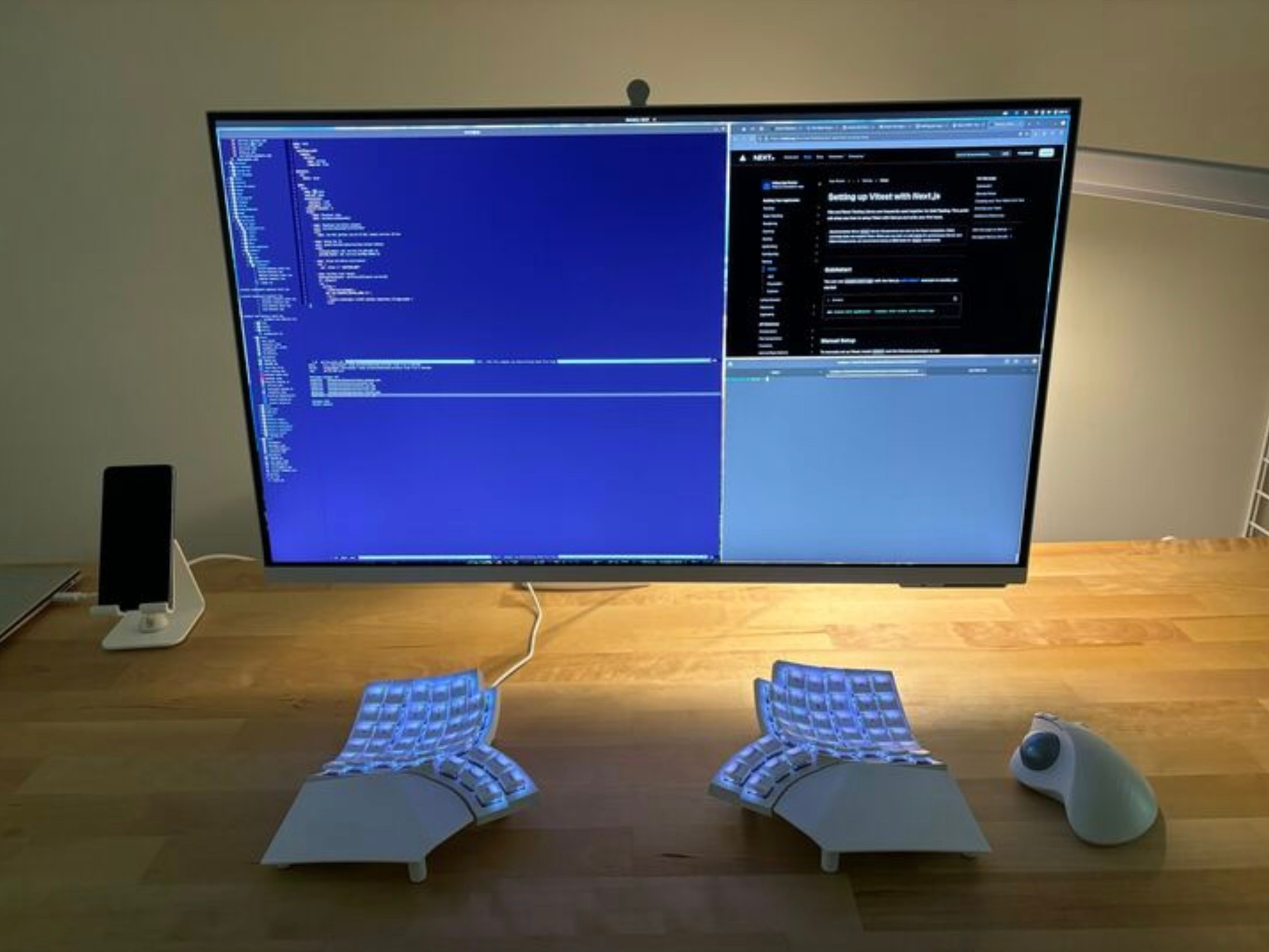 A minimalist coding desk setup featuring a large ultra-wide monitor displaying a split view with a blue terminal-based code editor on the left and the Arch Wiki website on the right, specifically a page about setting up Vite with React.js. Below the screen sits a white split ergonomic keyboard with backlit keys and a sculpted, tented layout. To the right is a white ergonomic trackball mouse, and to the left, a smartphone rests on a white stand. The entire setup is placed on a clean wooden desk with warm ambient lighting, emphasizing productivity and ergonomic design.