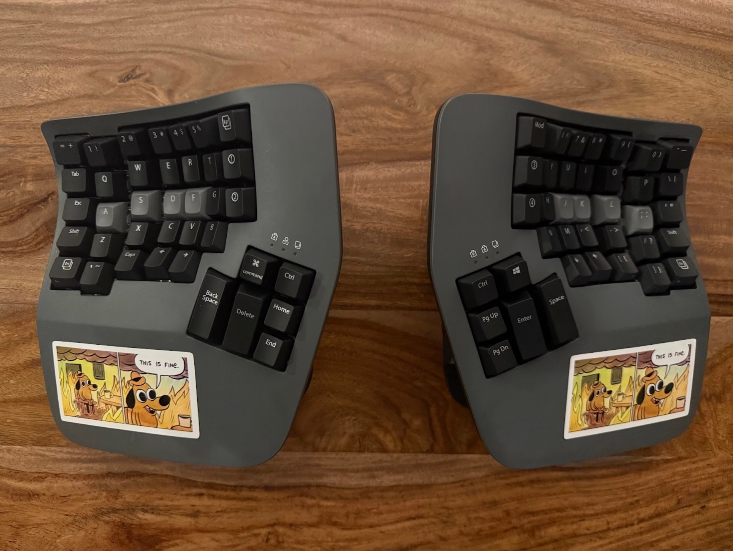 A split ergonomic keyboard with two separate dark-gray halves placed on a wooden surface. Each half features a column-staggered key layout with sculpted keycaps for improved typing ergonomics. The left half includes keys like “Backspace,” “Command,” and “Delete,” while the right half has “Enter,” “Space,” and “Ctrl.” Both halves have a sticker featuring the "This is fine" meme — a cartoon dog surrounded by flames calmly sipping coffee. The keyboard is customized for comfort and humor, appealing to enthusiasts and remote workers.