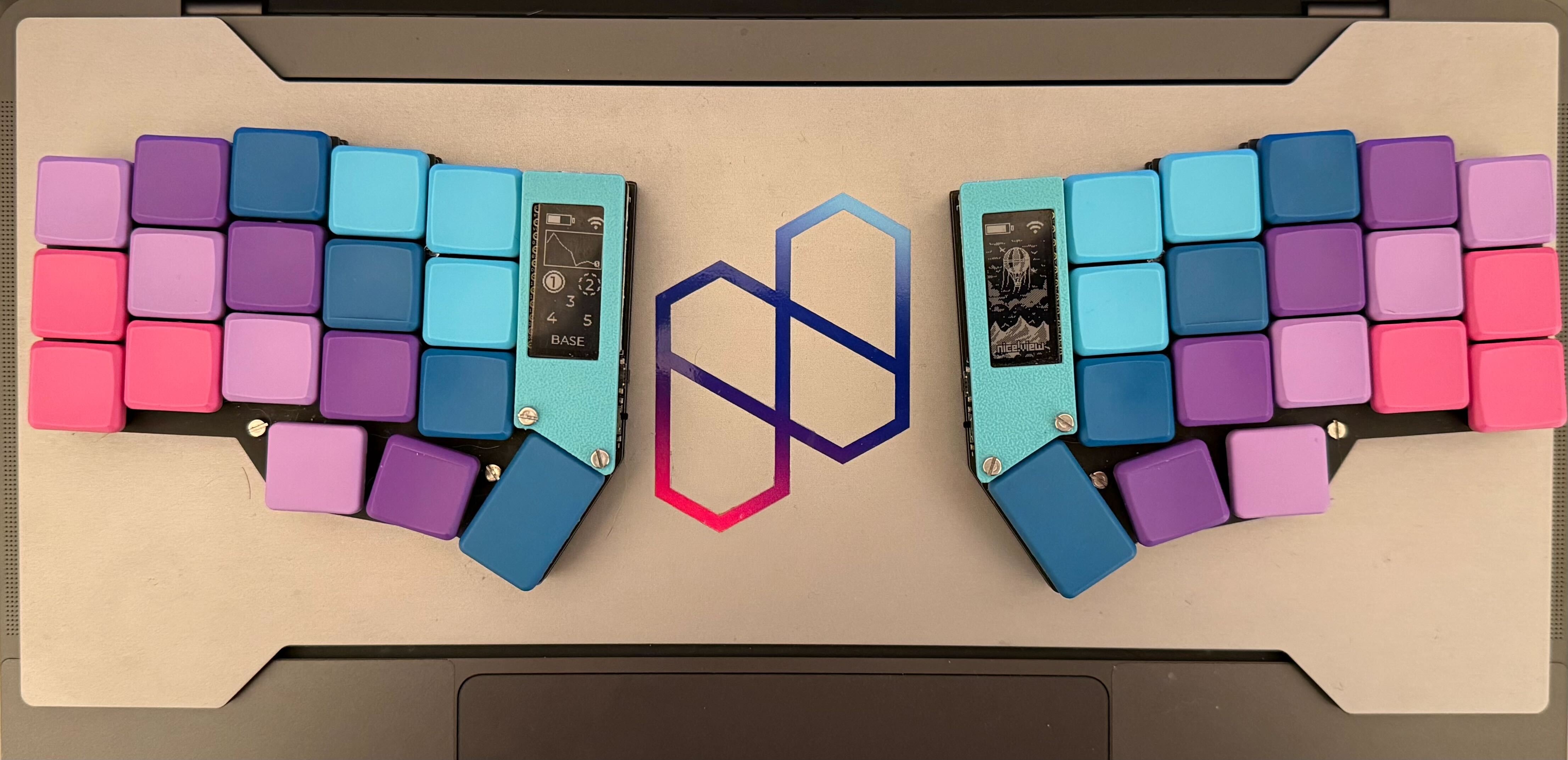A vibrant split ortholinear mechanical keyboard with pastel gradient keycaps ranging from pink and lavender to teal and deep purple. Each half of the keyboard is encased in a teal frame and connected by a flexible blue silicone bridge with a penguin logo in the center. The keyboard features built-in OLED displays; the left shows a layered mode interface labeled “BASE,” while the right shows a mountain and forest-themed graphic. The keyboard rests on a laptop with a colorful hexagonal logo beneath it.