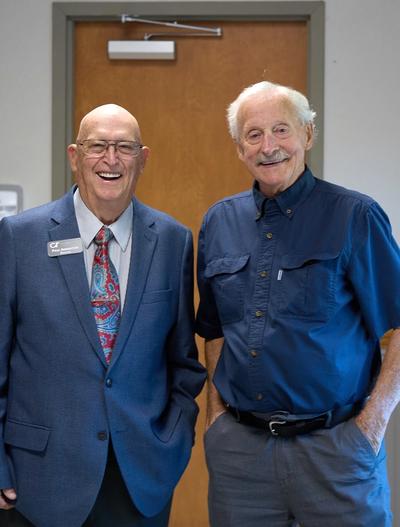 Two smiling senior men, one in a blue blazer and patterned tie, the other in a blue collared shirt.