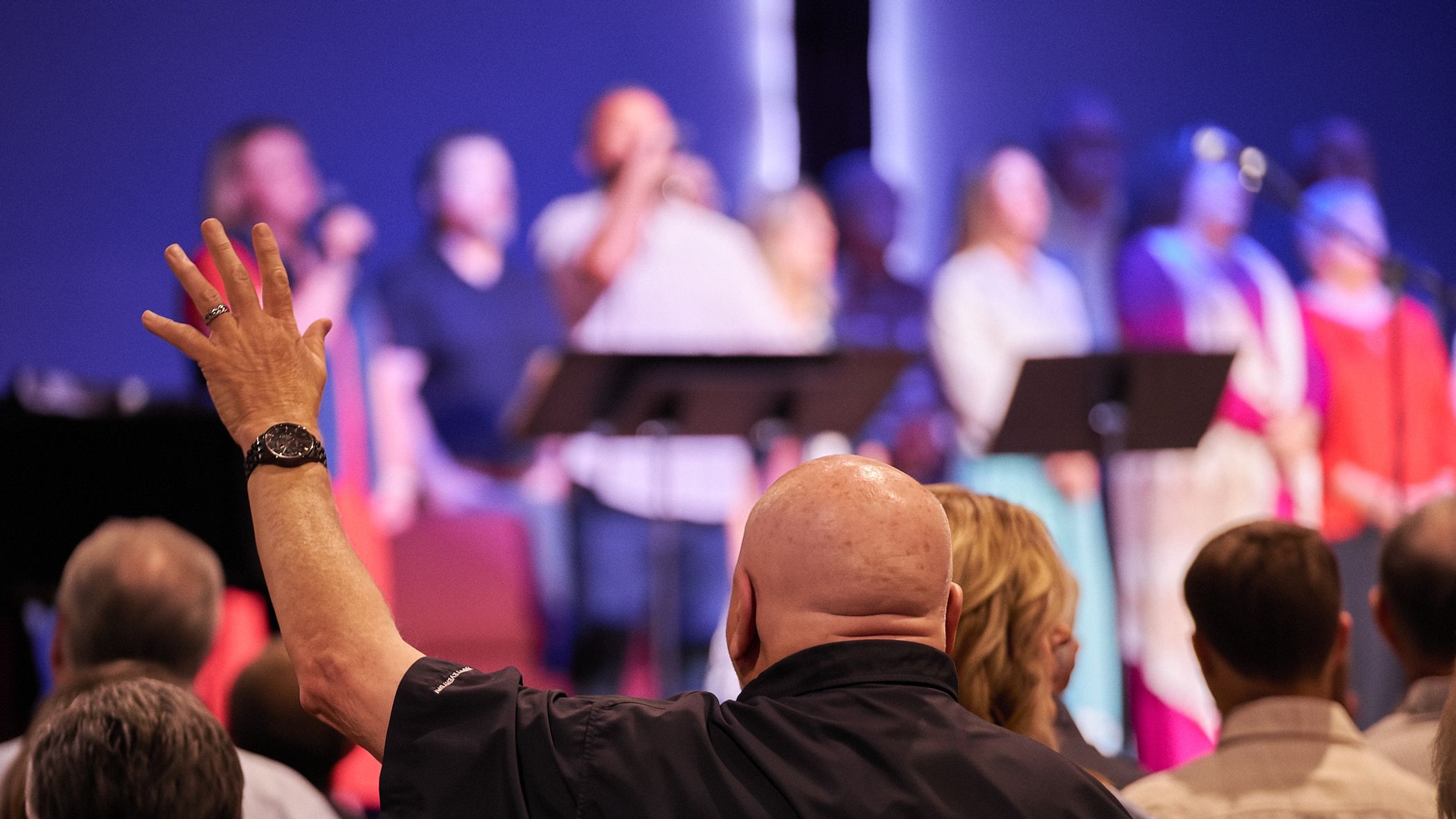 Person with a raised hand at a worship service, choir on stage in background.