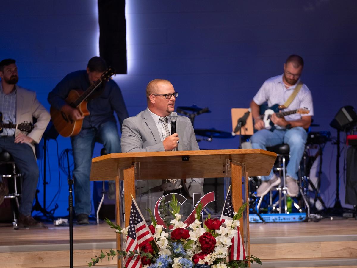 A man speaks at a podium decorated with American flags and flowers, while three men play guitars on a stage with a cross.