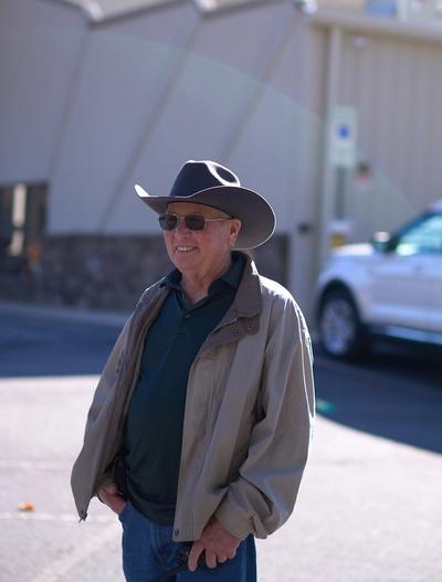 Smiling older man in a cowboy hat, sunglasses, and tan jacket holding a walkie-talkie.