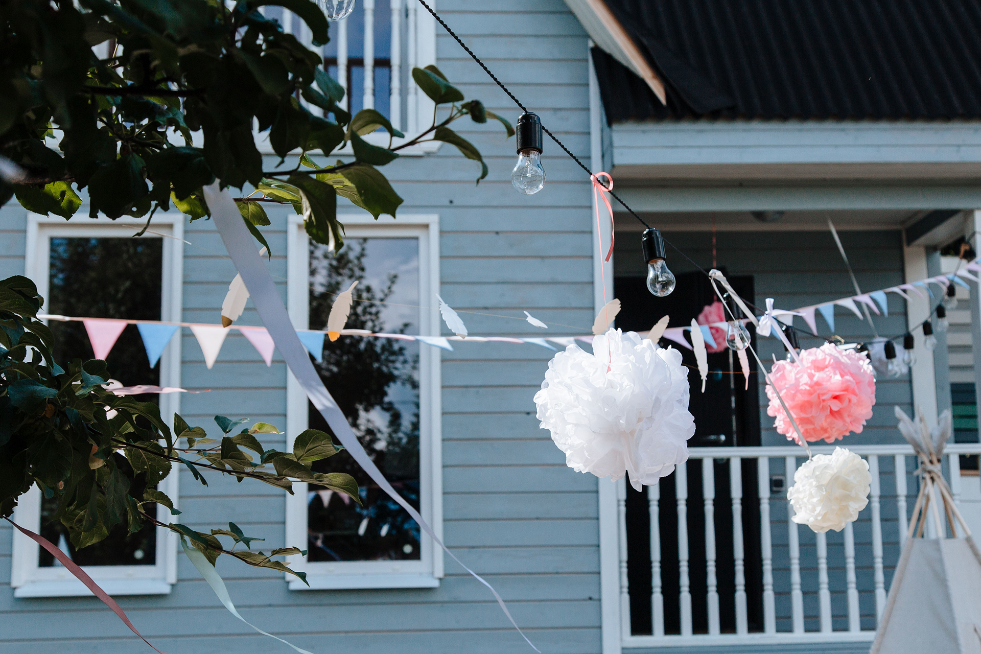 Elegant hanging decorations in an outdoor garden setting with pom poms and bunting against a wooden house.