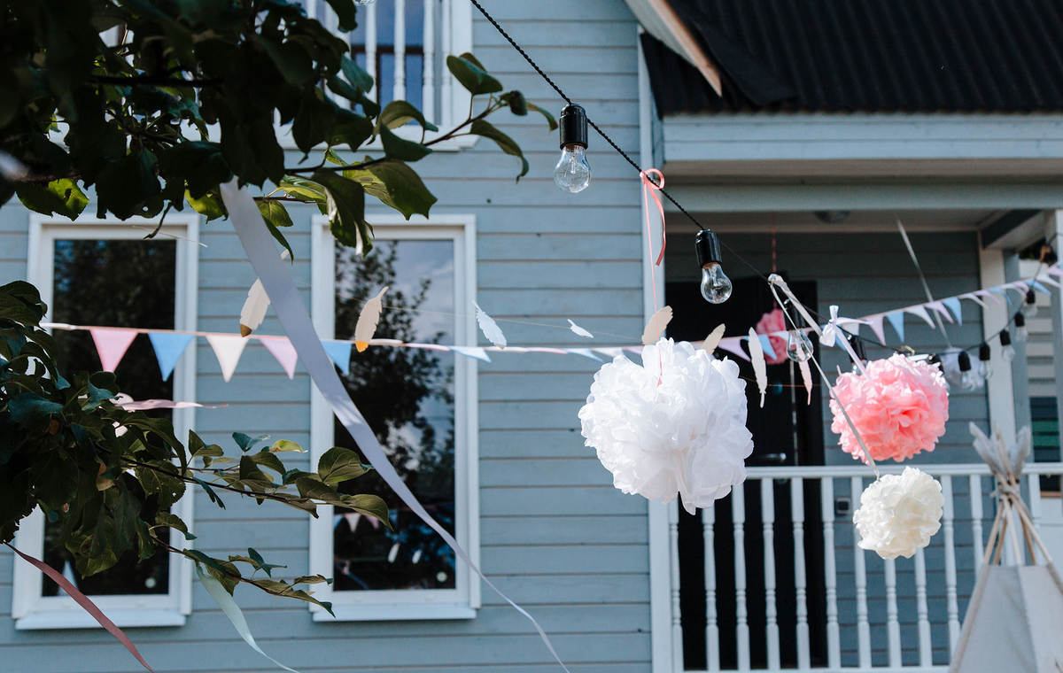 Elegant hanging decorations in an outdoor garden setting with pom poms and bunting against a wooden house.